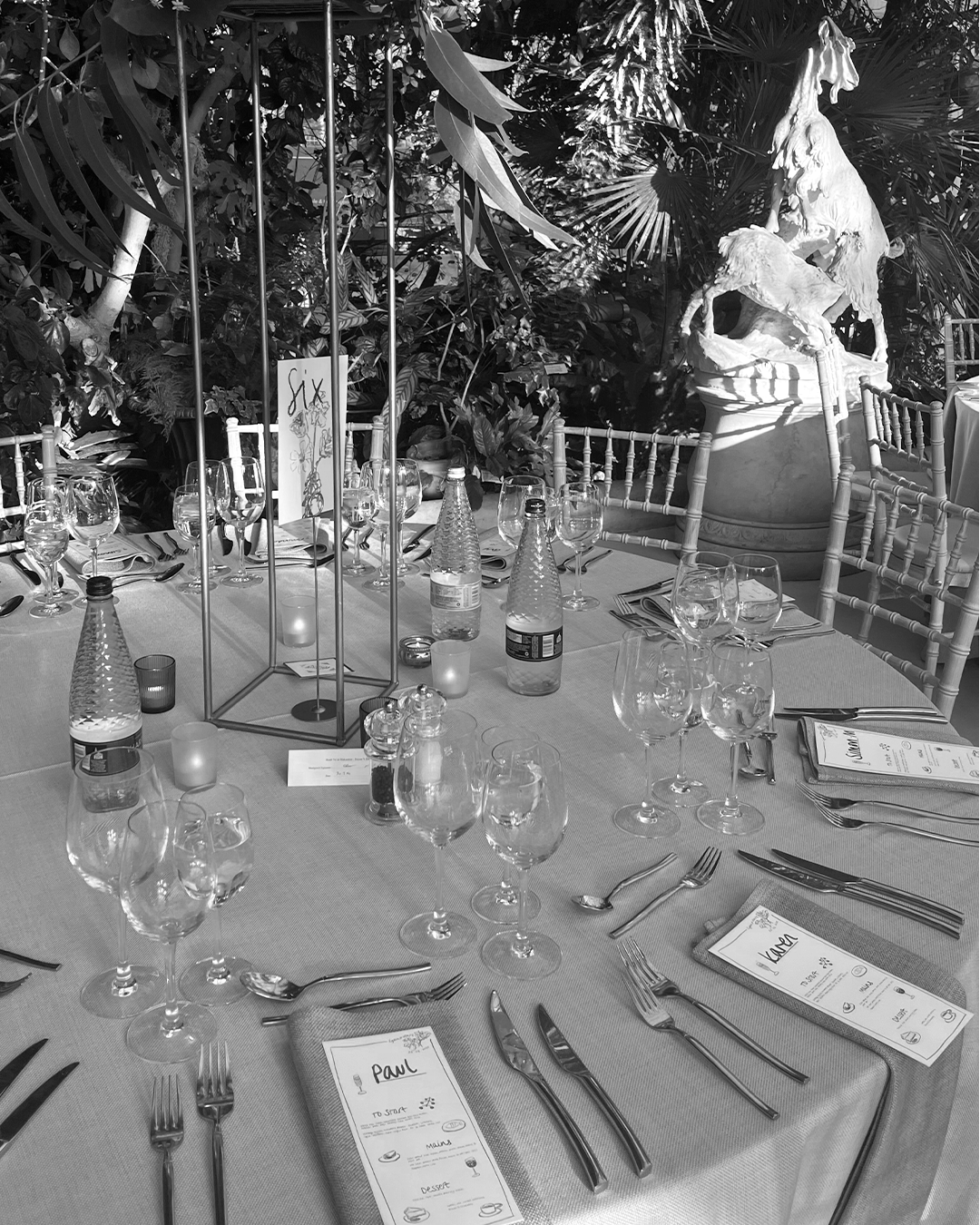 A banquet table set for a formal event with wine glasses, water bottles, menus labeled with names like Paul and Kevin, silverware, and candle lantern centerpieces. The background features a garden with lush foliage and a statue of a horse.