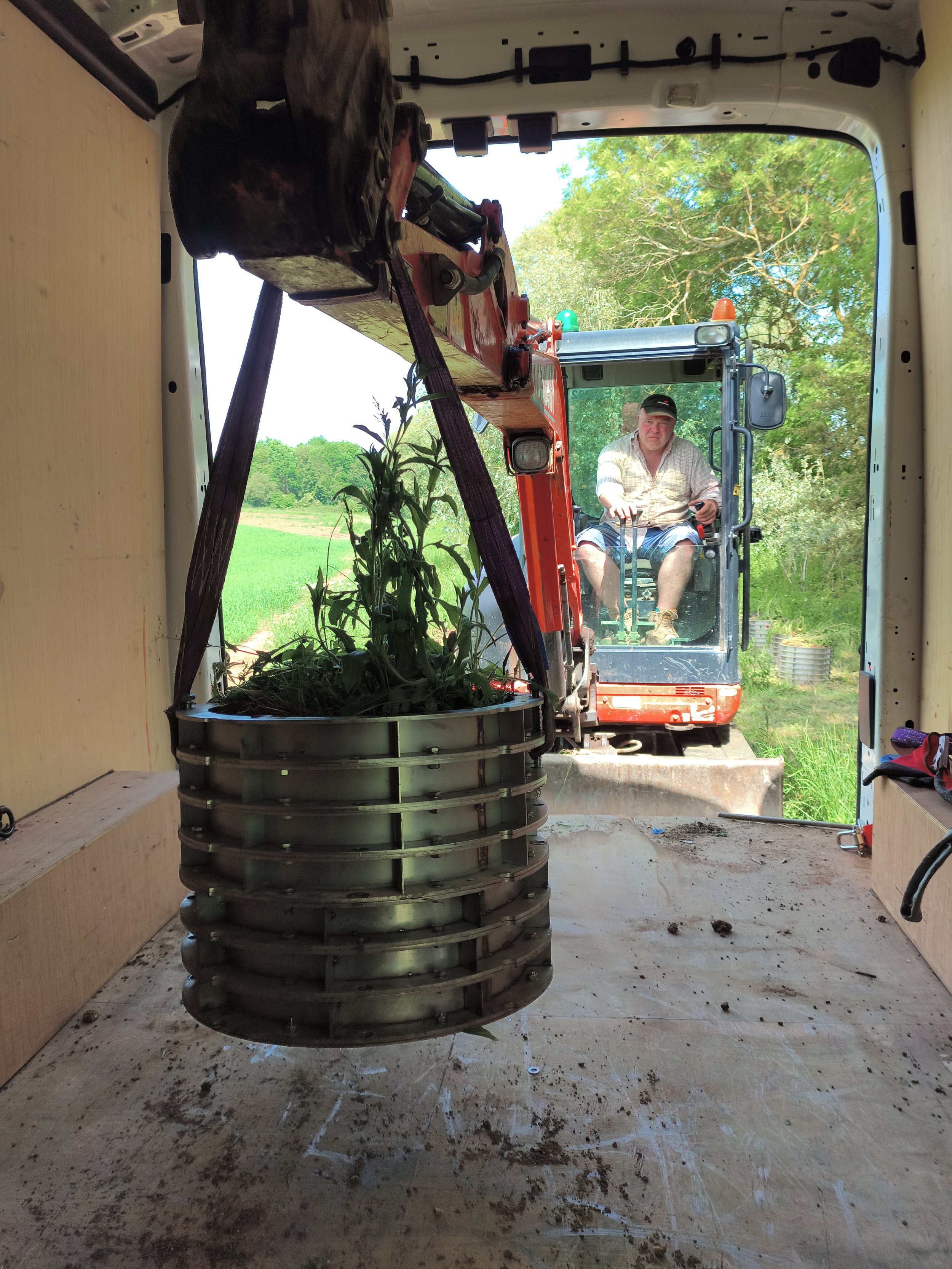 Inside a truck, a vertical metal tumbler with plants growing inside is in the foreground. The view is from inside looking out through a doorway with a tractor and person visible outside in a rural setting with green trees and fields.