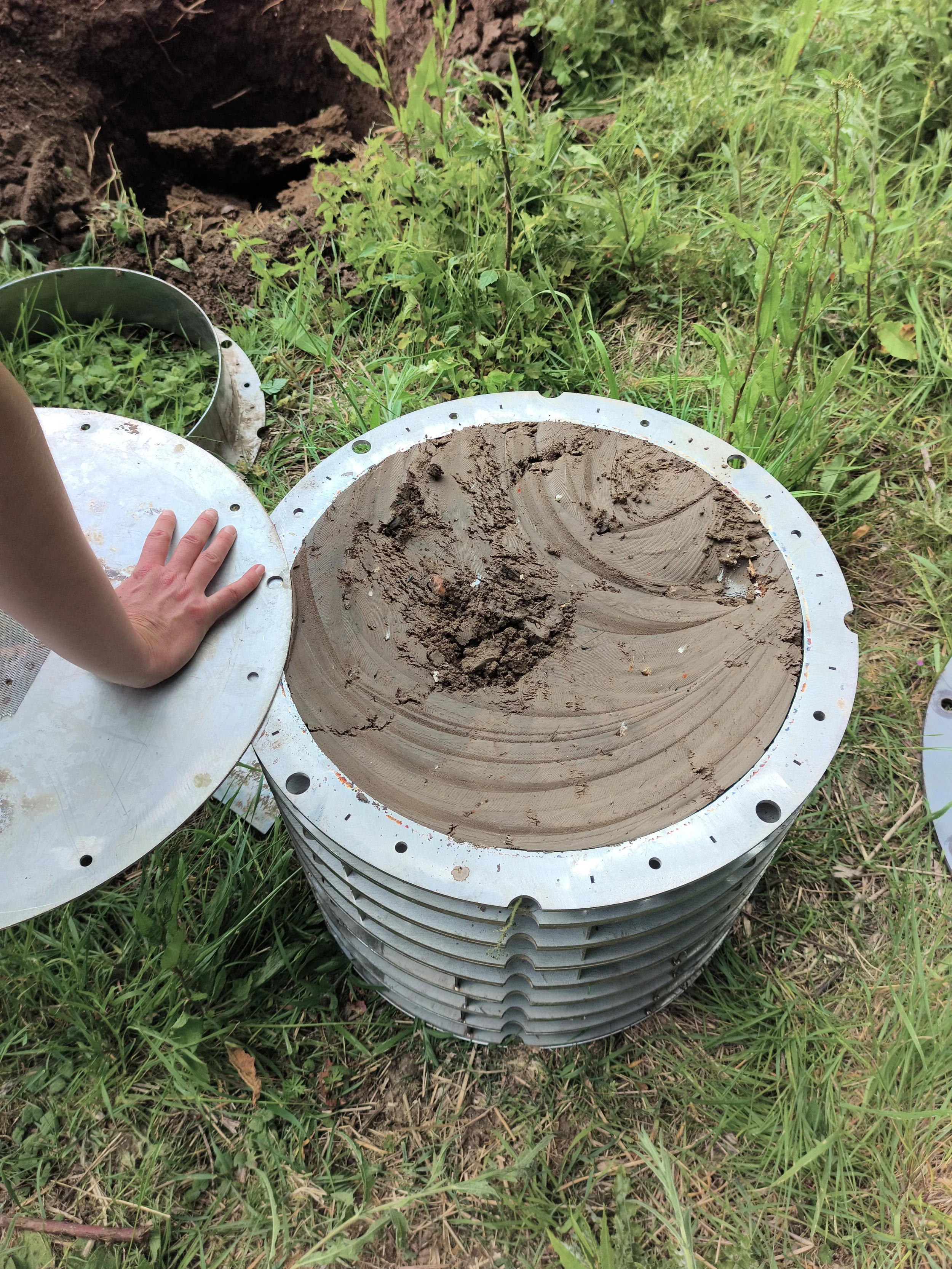 Stacked metal parts with dirt and grass on top, near a hole in the ground in an outdoor grassy area.
