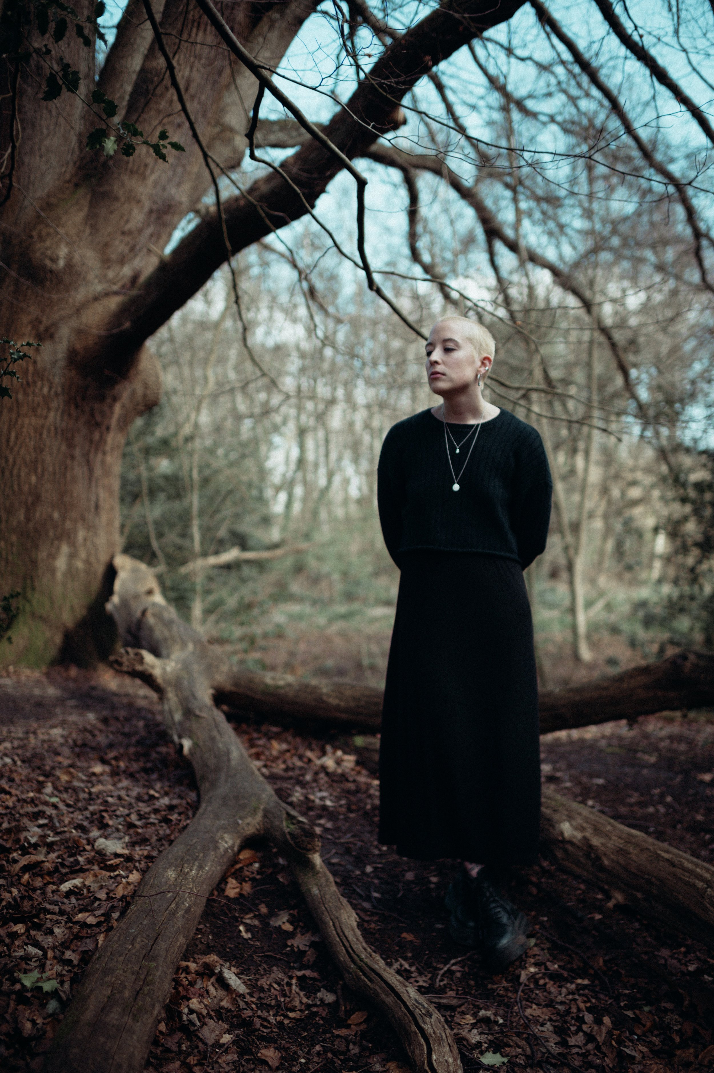 A person with short blonde hair, wearing a black sweater and layered necklaces, standing outdoors in a wooded area with leafless trees and fallen leaves, near a large fallen tree branch.