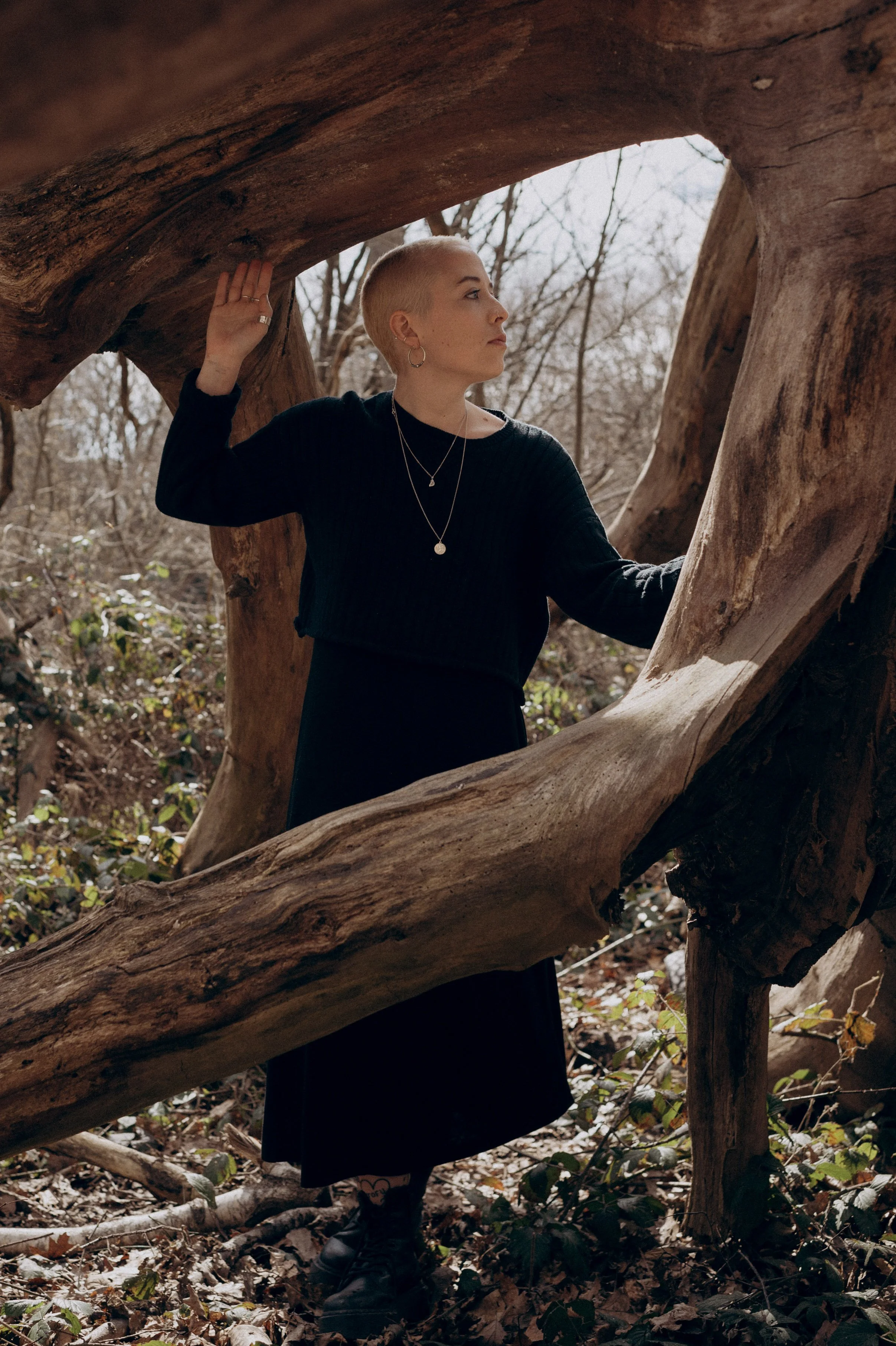 A person with short blonde hair and multiple earrings is standing outdoors in a wooded area, near a large fallen tree. They are wearing a black dress or top, multiple necklaces, and black combat boots, and are holding onto the tree with one hand whil