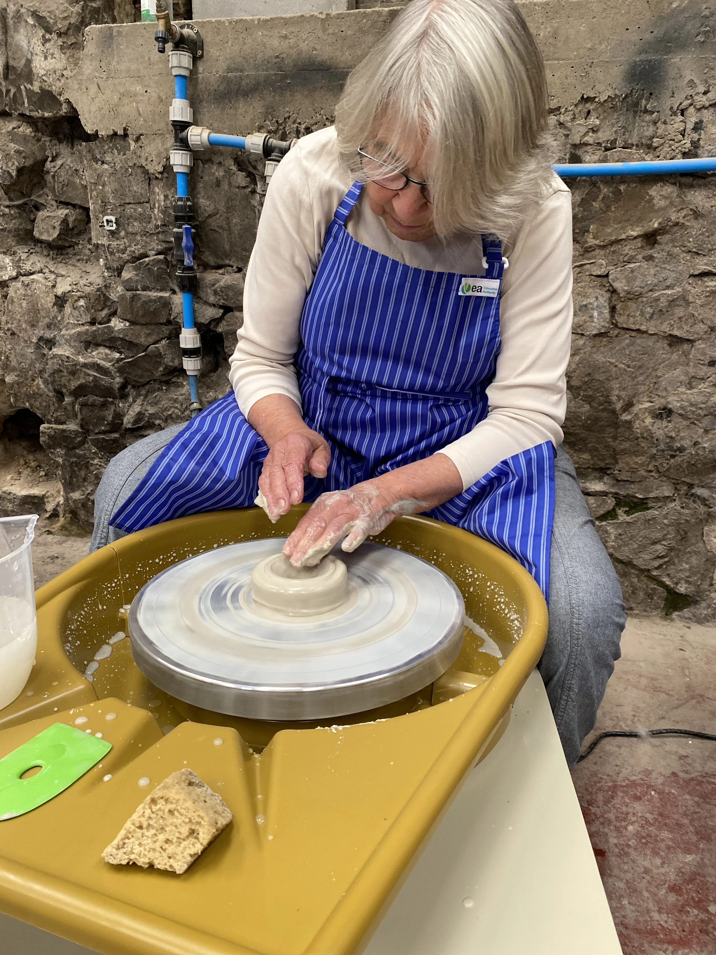 A woman in jeans, cream jumper and a blue apron working at a potters wheel in an old stone bard