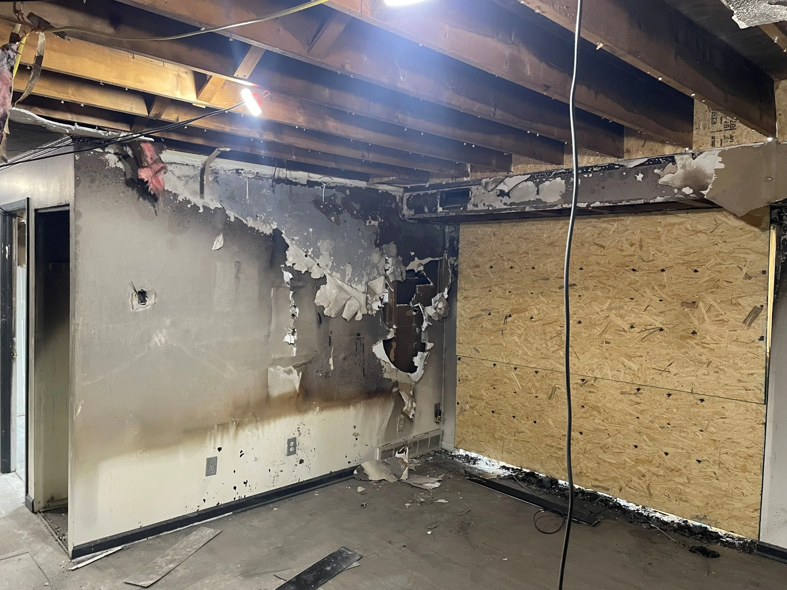 Partially burned and damaged interior wall with electrical outlets, charred and peeling wall paint, and a large plywood sheet covering a damaged section, with exposed ceiling beams and wiring.  Significant aftermath of fire and smoke damage with soot