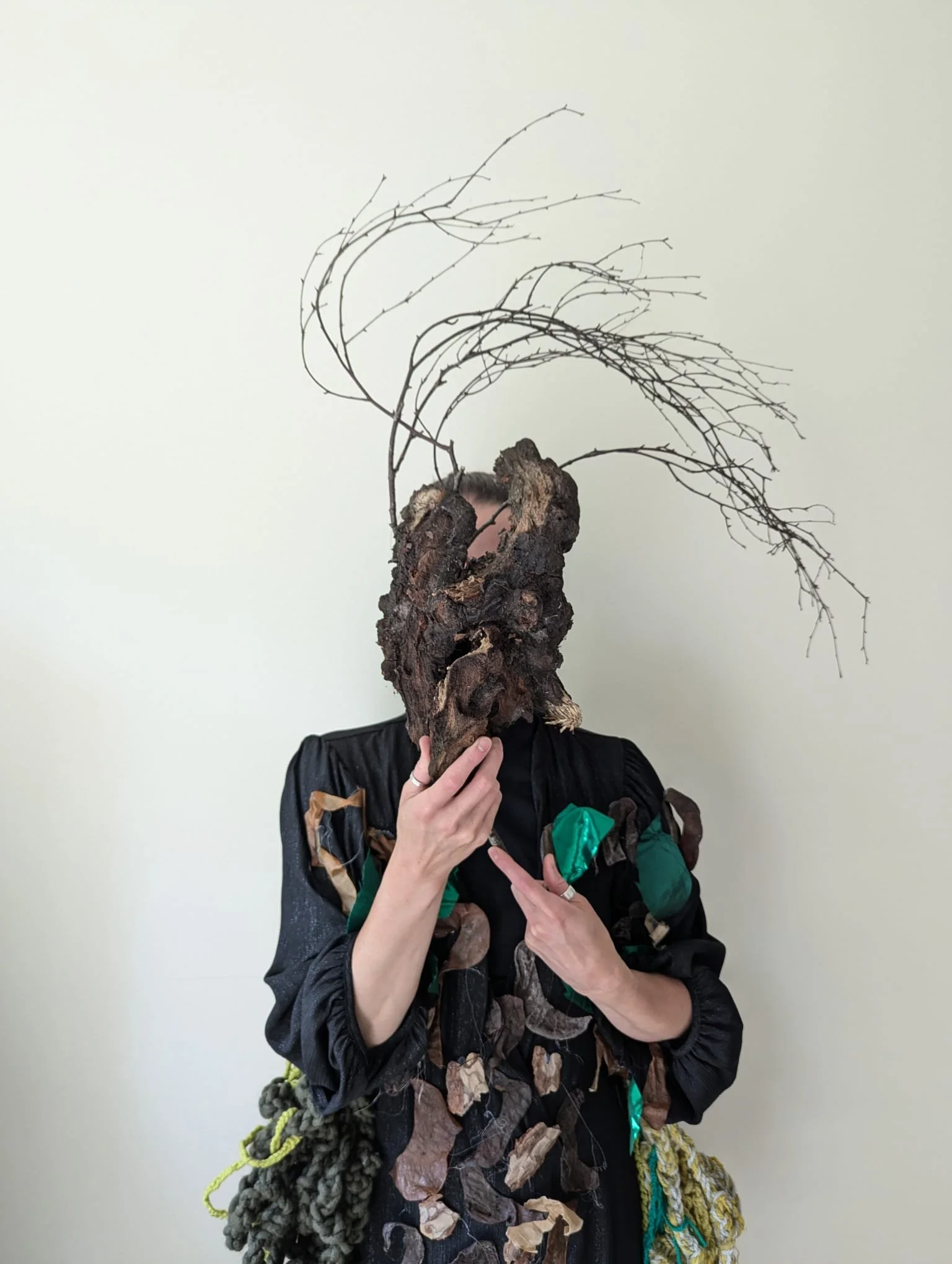 © Judita Vivas
An image of a performer dressed in a patchy green, brown and back costume, covering her face with a piece of wood with dried, thin tree branches sticking up, making it resemble a mask.