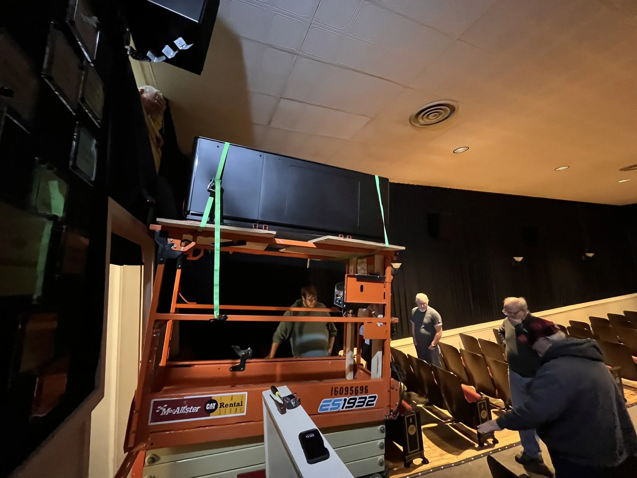 Several people are loading the new projector into the projection booth on an orange lift inside the auditorium.