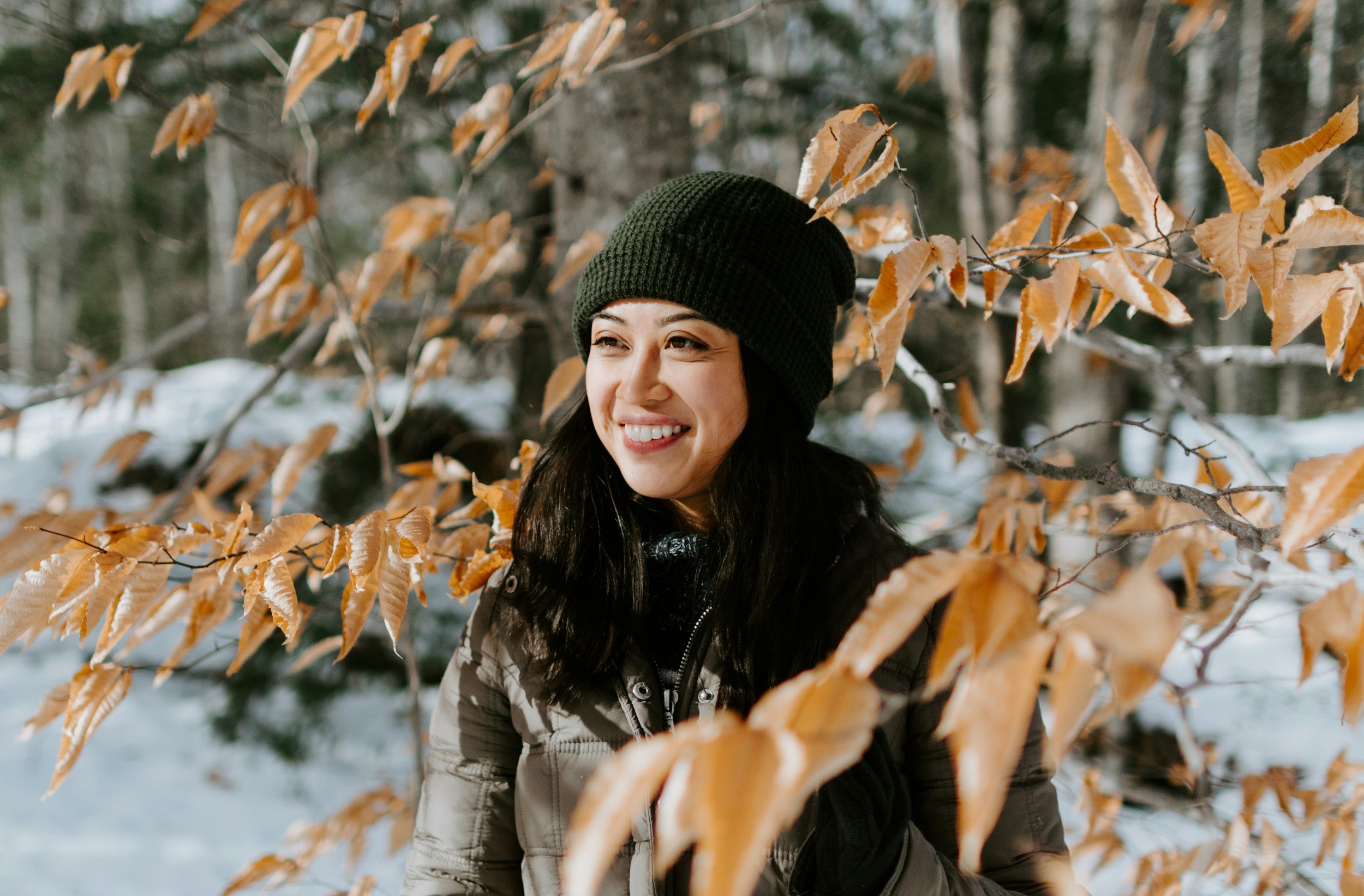 A woman smiling in a winter forest with orange leaves on trees around her, wearing a green beanie and a beige jacket.