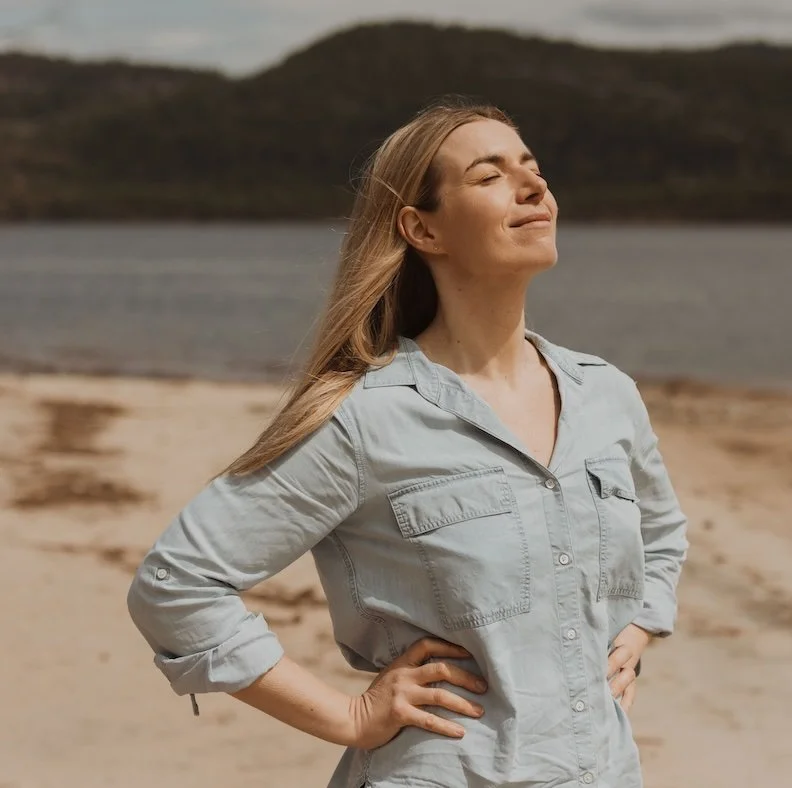 A woman with long blonde hair standing on a beach with her eyes closed and a content smile, wearing a light gray button-up shirt with rolled-up sleeves, with water and hills in the background.