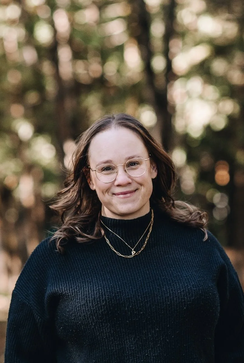 A woman with shoulder-length brown hair, glasses, and gold jewelry smiling outdoors in front of trees during daytime.