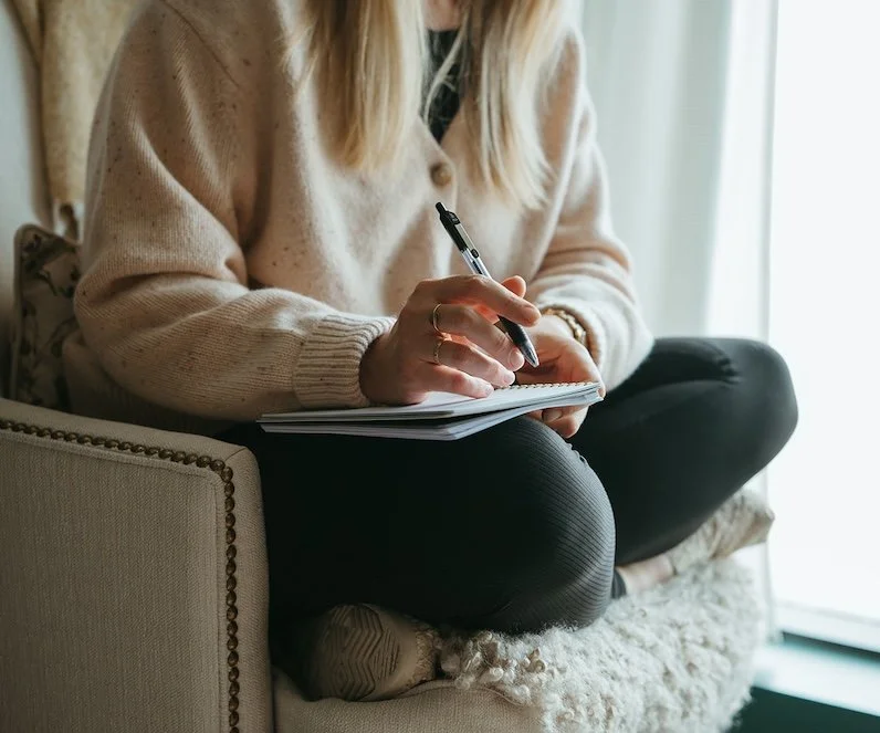 Person sitting on a cushioned chair, writing in a notepad with a pen, wearing a beige sweater and black pants.