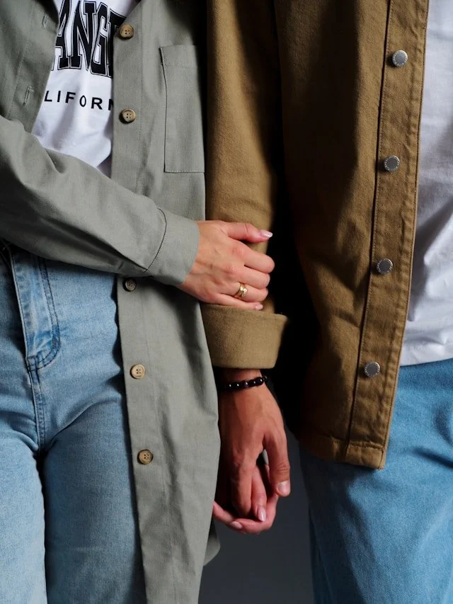 Two people holding hands, with one wearing a light green jacket and the other in a brown jacket, both wearing jeans.