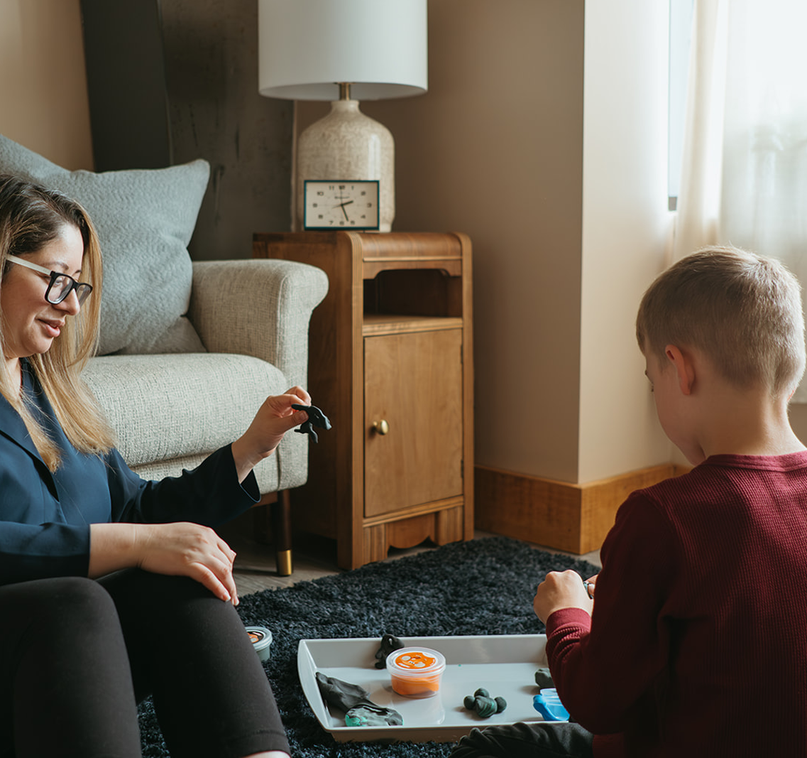 A female therapist and a boy play with modelling clay while sitting on a dark blue shag rug in a therapy office.