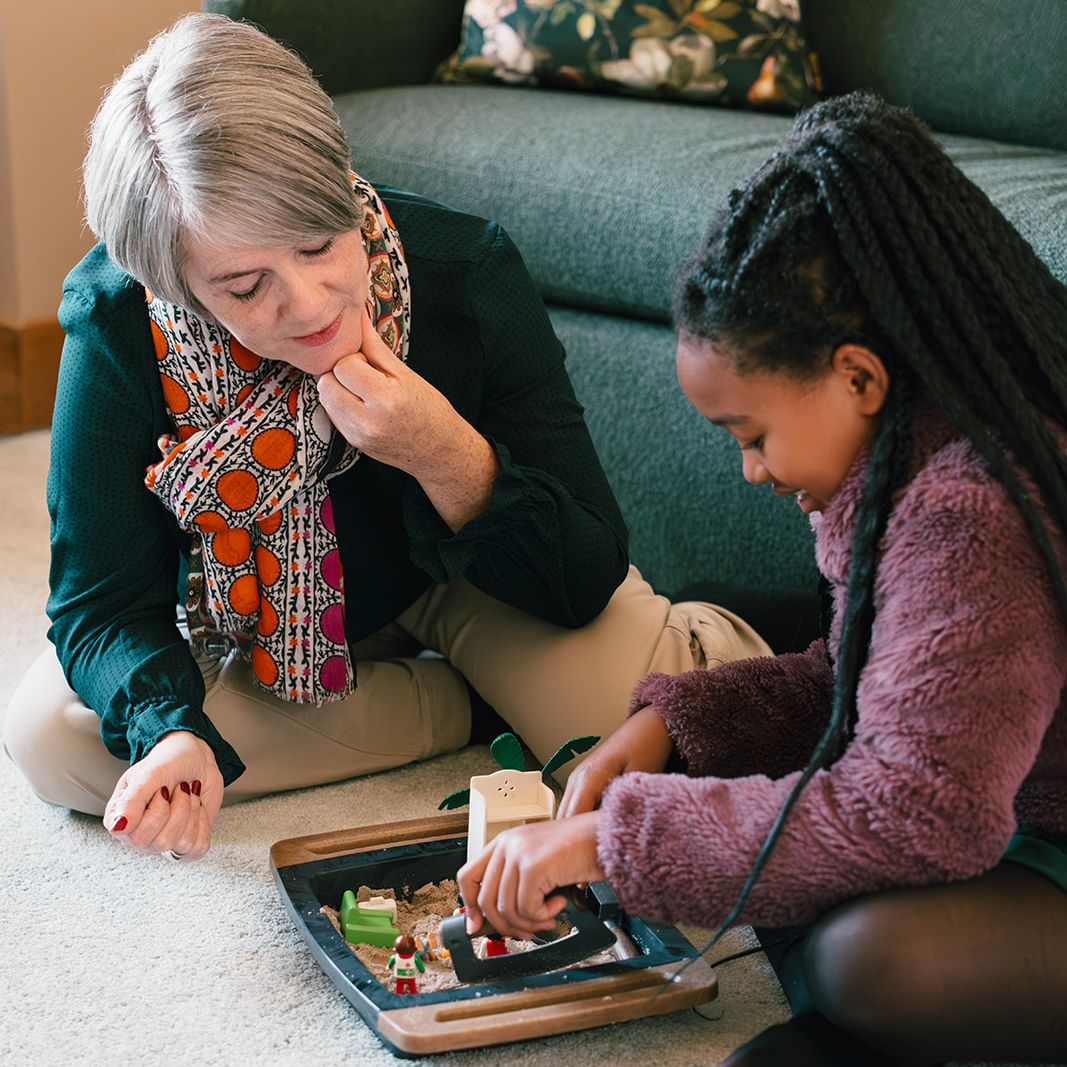 A female therapist and a young girl play with toys in a sand tray on a carpeted floor in a therapy office.