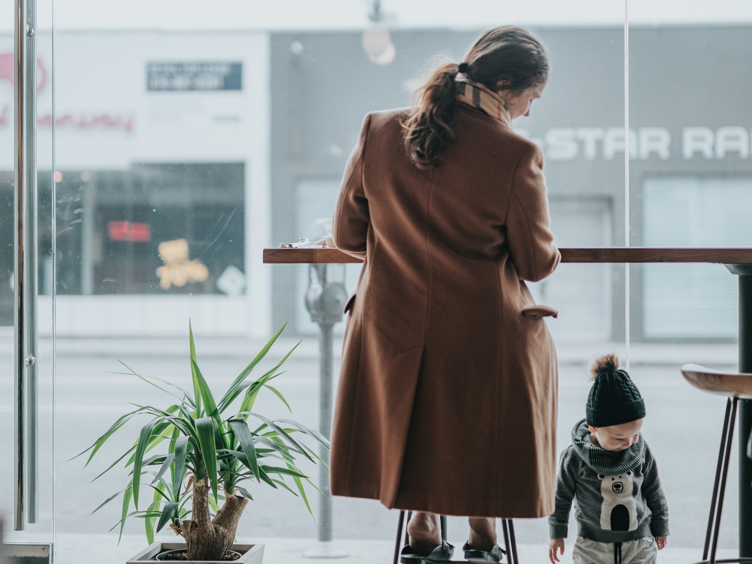 A woman wearing a scarf and coat looks down at a young child wearing a hat, scarf and sweater. She is sitting at raised table in a cafe and the child looks like it may go exploring any moment.