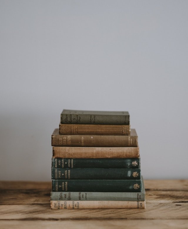 Stack of nine old hardcover books on a wooden surface with a plain gray wall in the background.