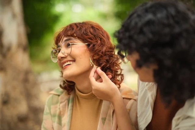 A woman with curly red hair and glasses smiling as she is helped with an earring by a person with dark hair outdoors in a green, forested setting.