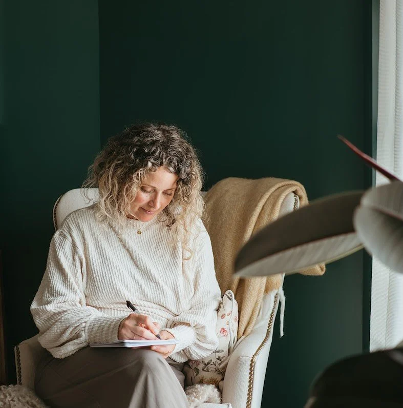 A therapist with curly hair sits on a beige armchair, writing in a notepad with a pen, in a room with dark green walls and a window with white curtains.