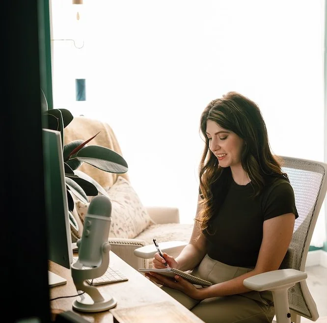 A female therapists sits at a desk, smiling while taking notes with a pen in a notebook, in front of a computer, with indoor plants nearby and bright natural light coming from a window.
