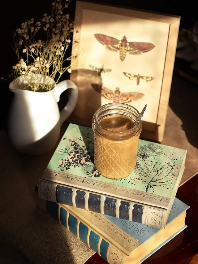 A cozy scene with a white pitcher holding dried flowers, a framed illustration of moths, a jar of honey with a spoon, and three stacked decorative books with floral and botanical covers.