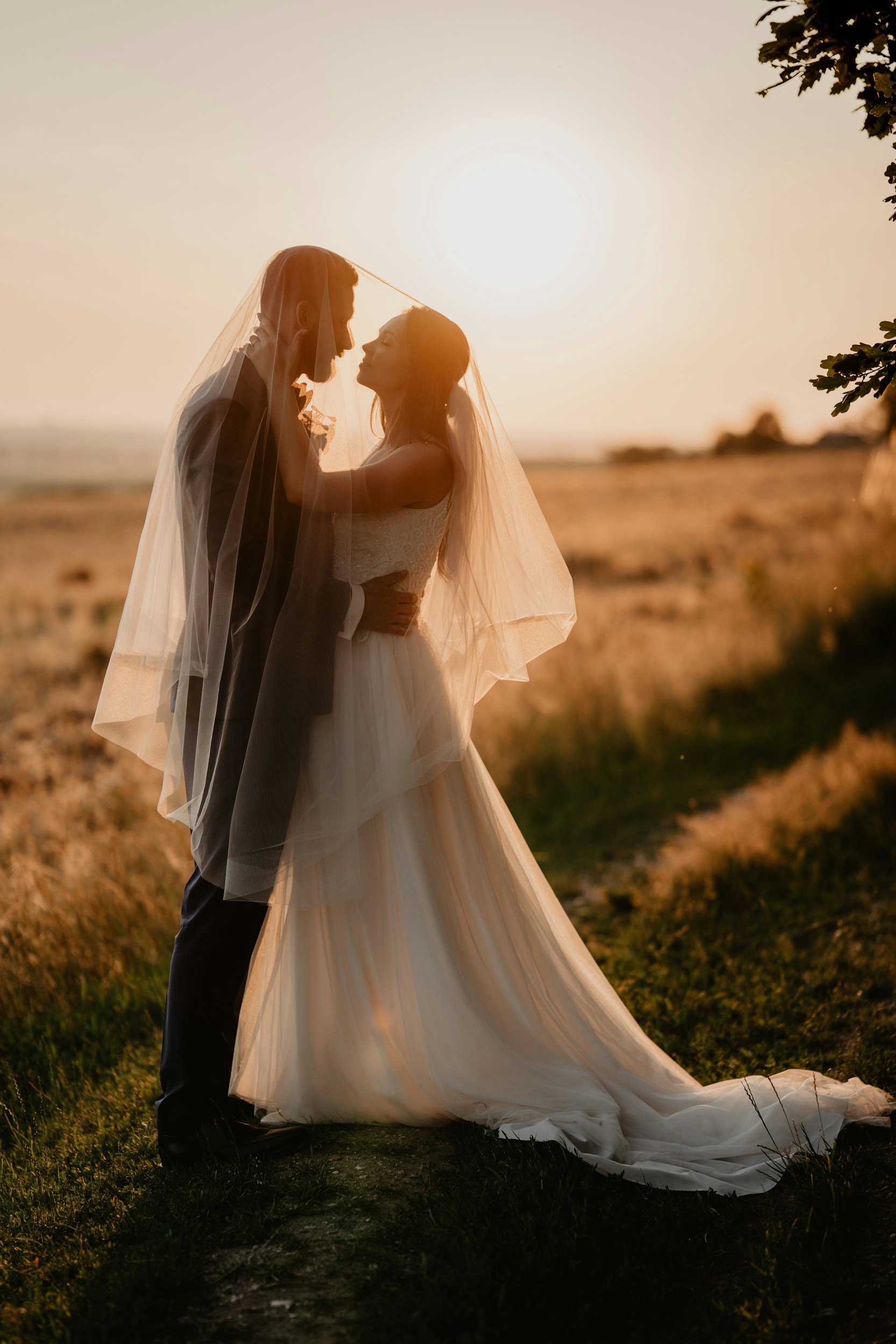 Bride and groom embracing under veil at sunset in a field.