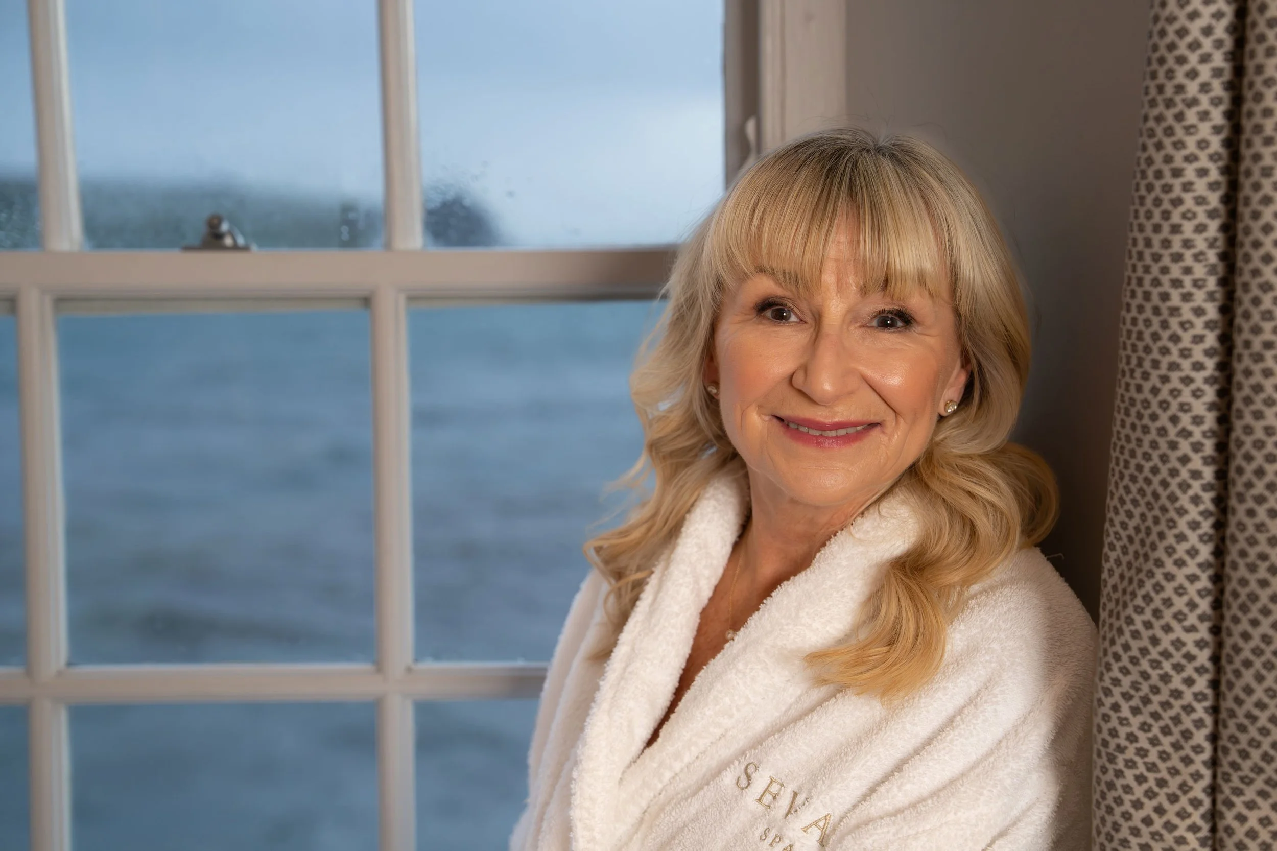 A smiling woman with blonde hair and a white towel with 'SEVAN' embroidered on it, sitting in front of a window with a view over the sea in St Mawes.
