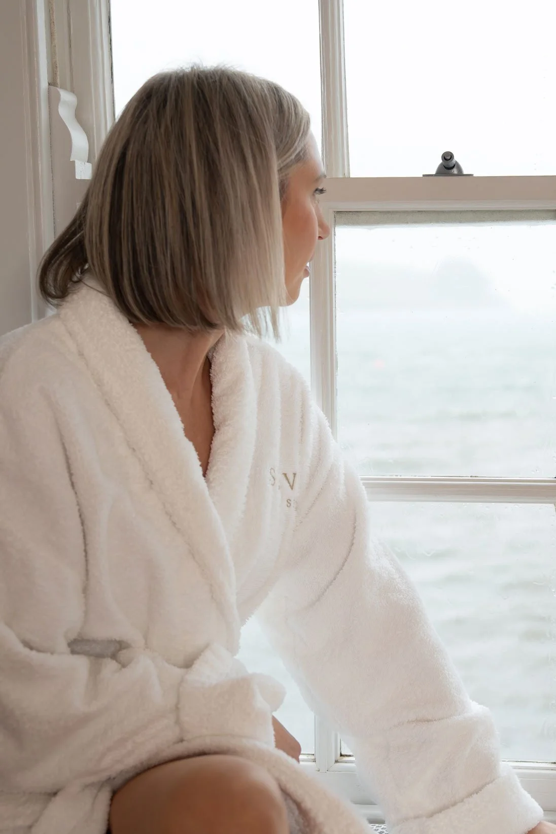 A woman with blonde hair looking out of a window, wearing a white bathrobe with initials embroidered on it, sitting near a body of water.