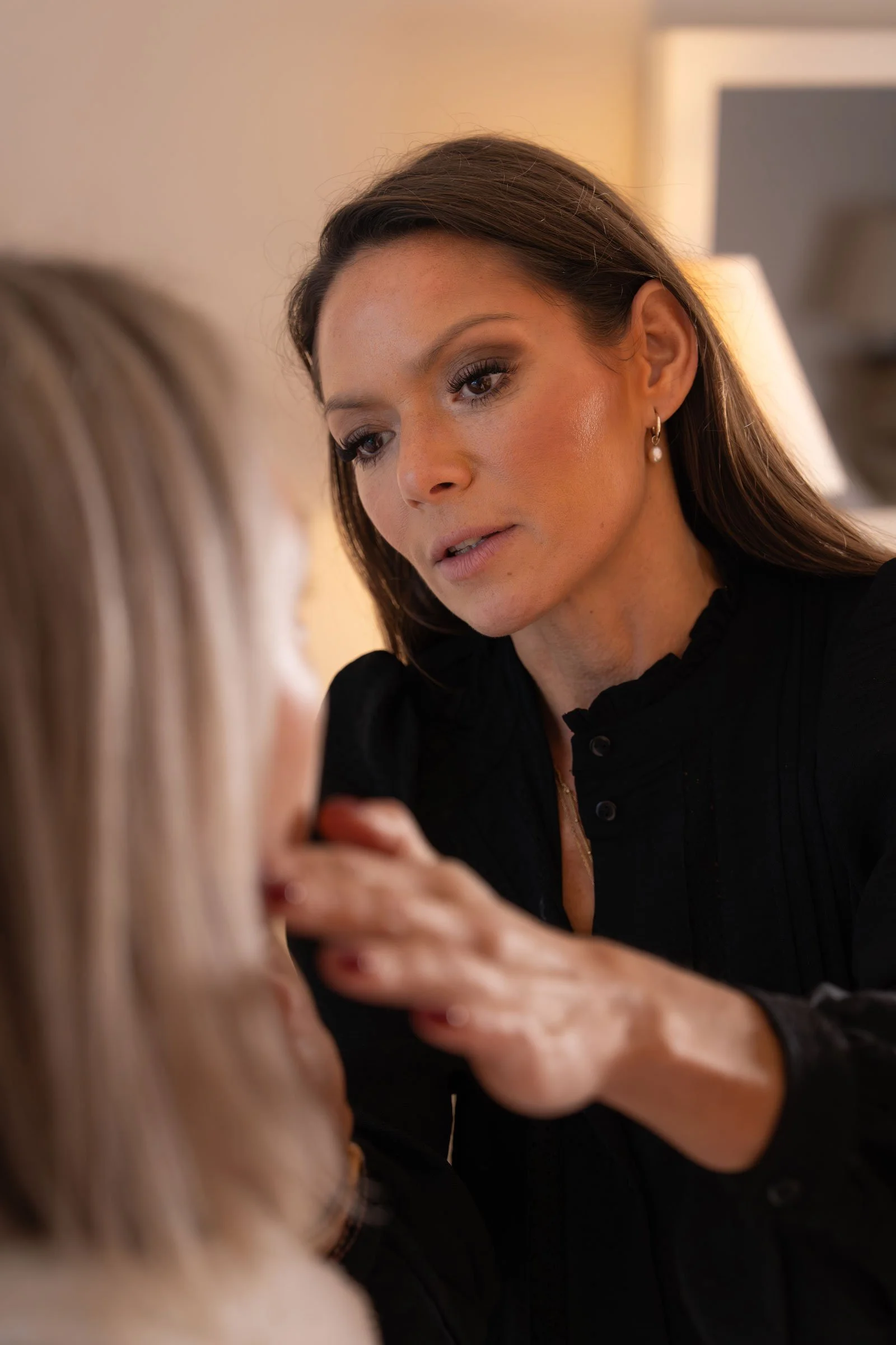A woman with brown hair and earrings is applying makeup to another woman, who has blonde hair, in a softly lit room.