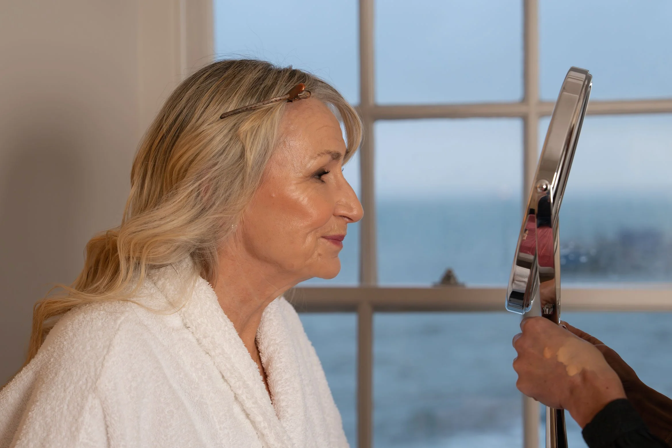 Woman in her 60s with blonde hair, wearing a white robe, looking into a mirror held by another person, with a window and view of the sea in the background.