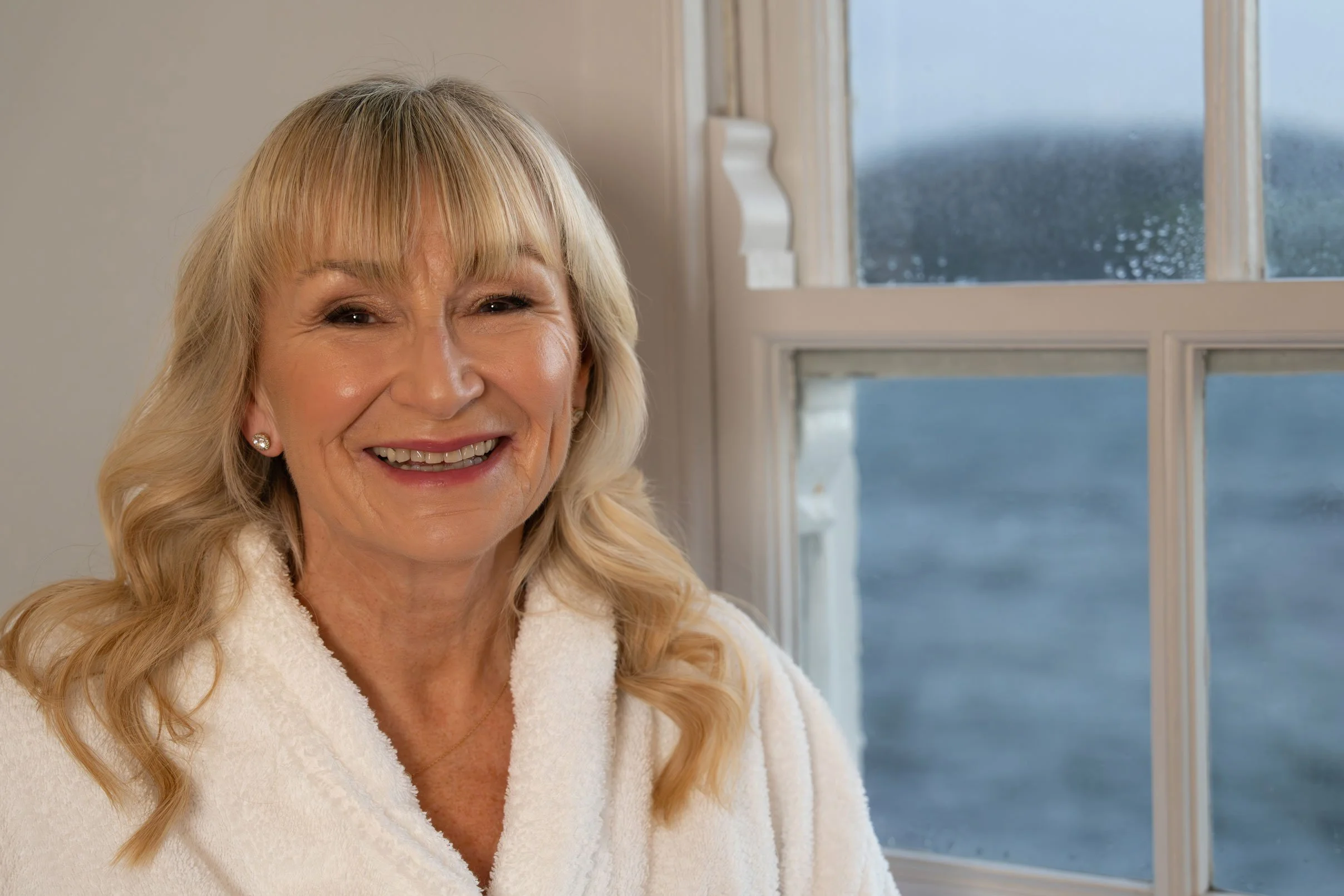 A smiling woman with blonde hair and earrings, wearing a white robe, sitting by a sash window view a view of the sea.