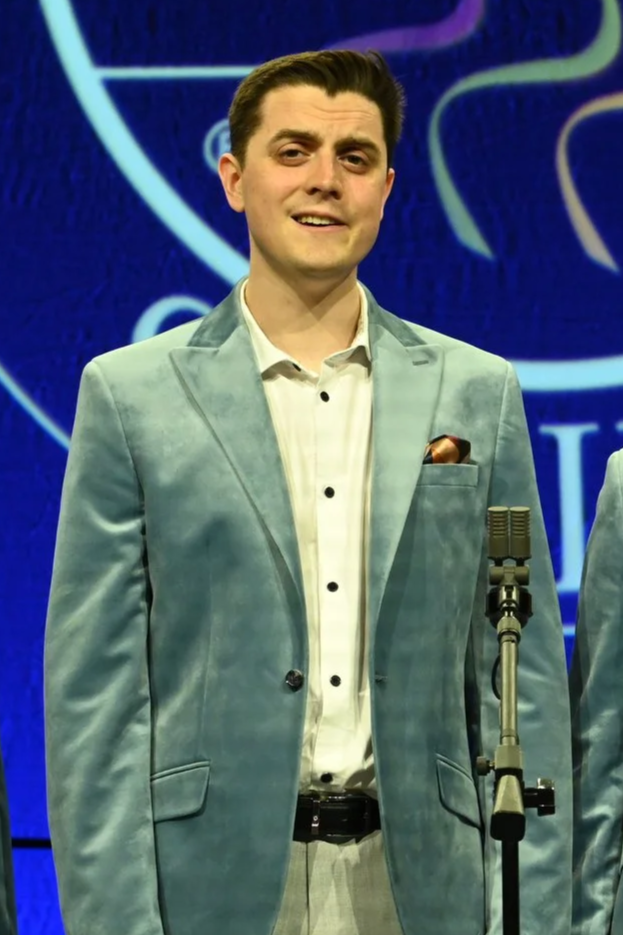 Oli Goodall suit standing in front of a microphone on a stage, with the Barbershop Harmony Society logo background.