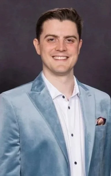 Oli Goodall wearing a light blue velvet blazer and a white shirt, against a dark background, smiling.