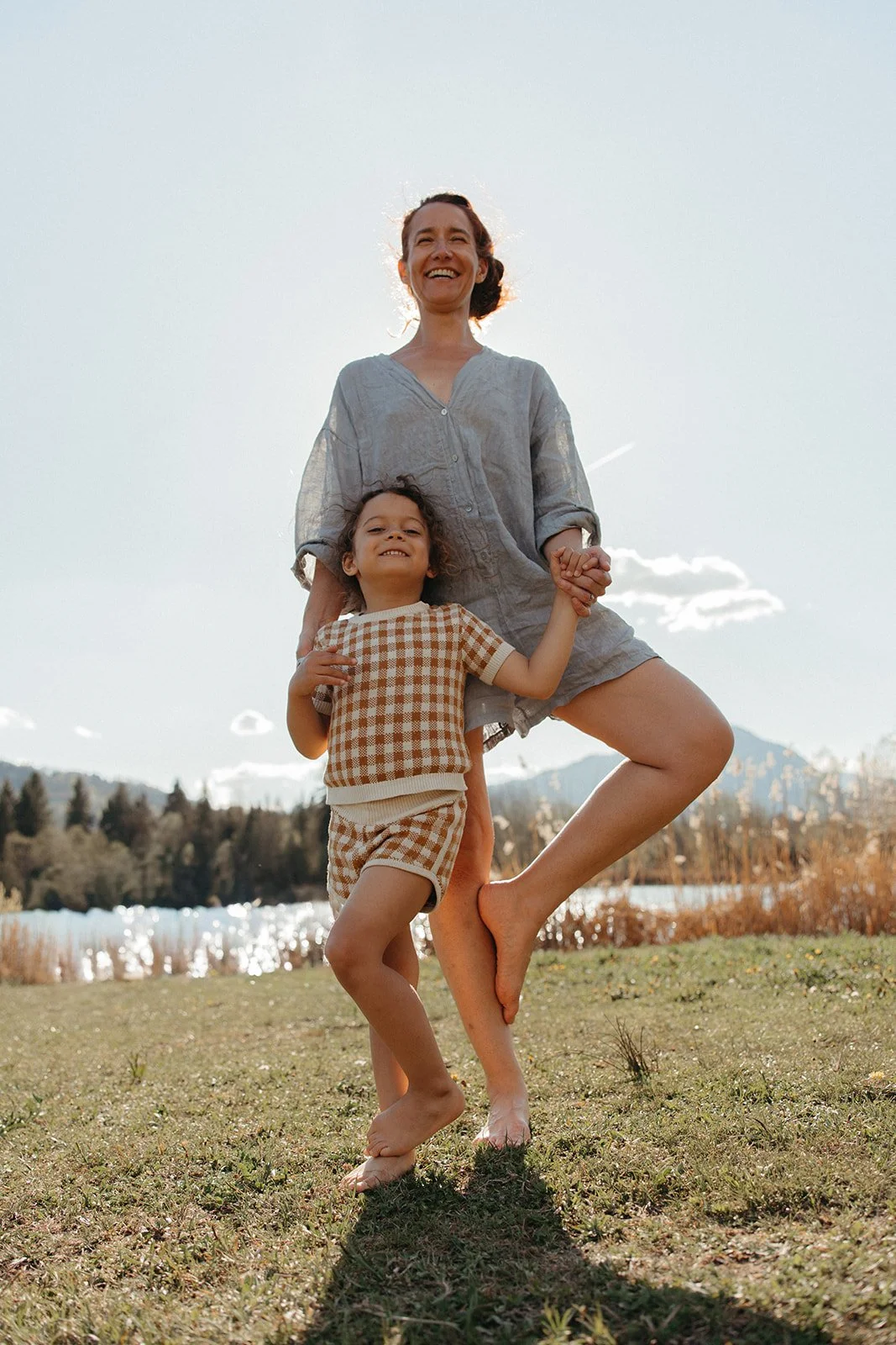 A woman and a young girl standing outdoors in a grassy area with a lake, trees, and mountains in the background, smiling and holding hands.