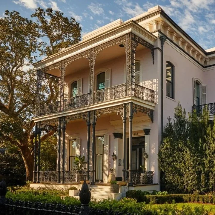 A large, white, two-story house featuring ornate black wrought-iron balconies on both floors, tall windows with shutters, and a front porch with pillars. Design by Olivia Erwin Interiors featured in Architectural Digest.