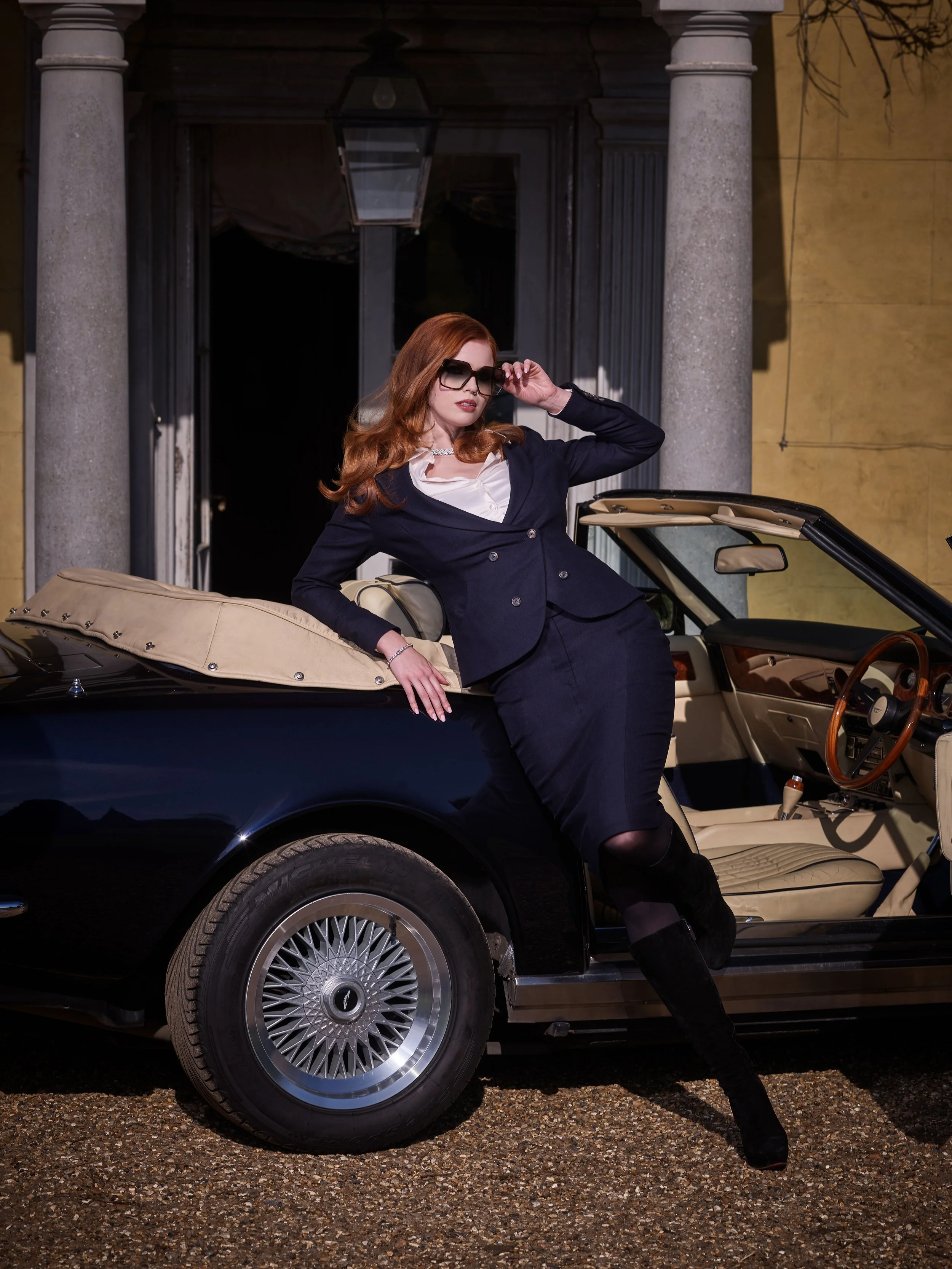 British luxury womenswear editorial featuring a navy tailored skirt suit consisting of a pencil skirt and blazer styled with knee-high boots beside a classic convertible car.