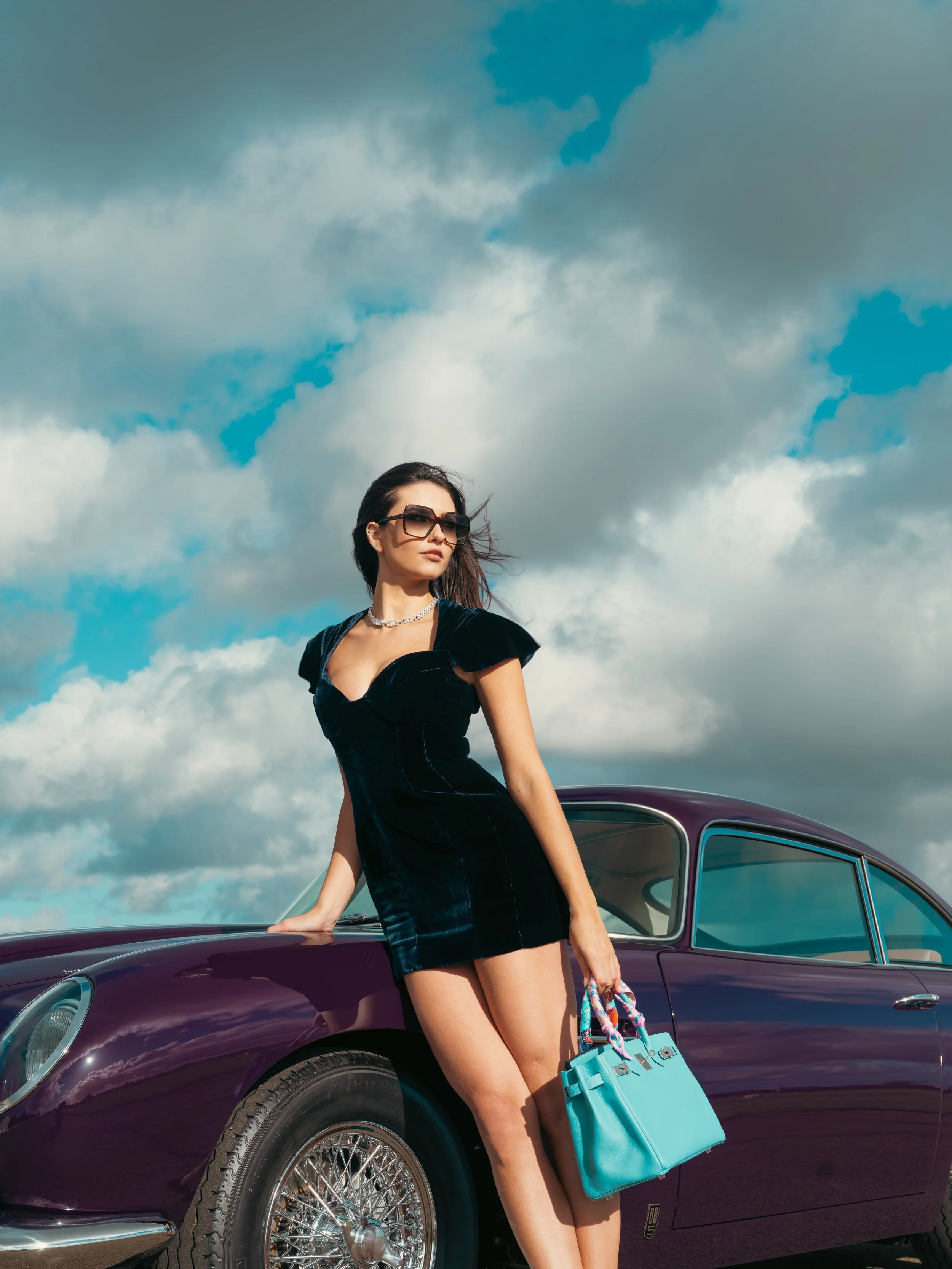 Navy velvet corset midi dress by Paget London, styled standing beside a classic car and paired with a Hermès Birkin bag in a luxury fashion editorial.