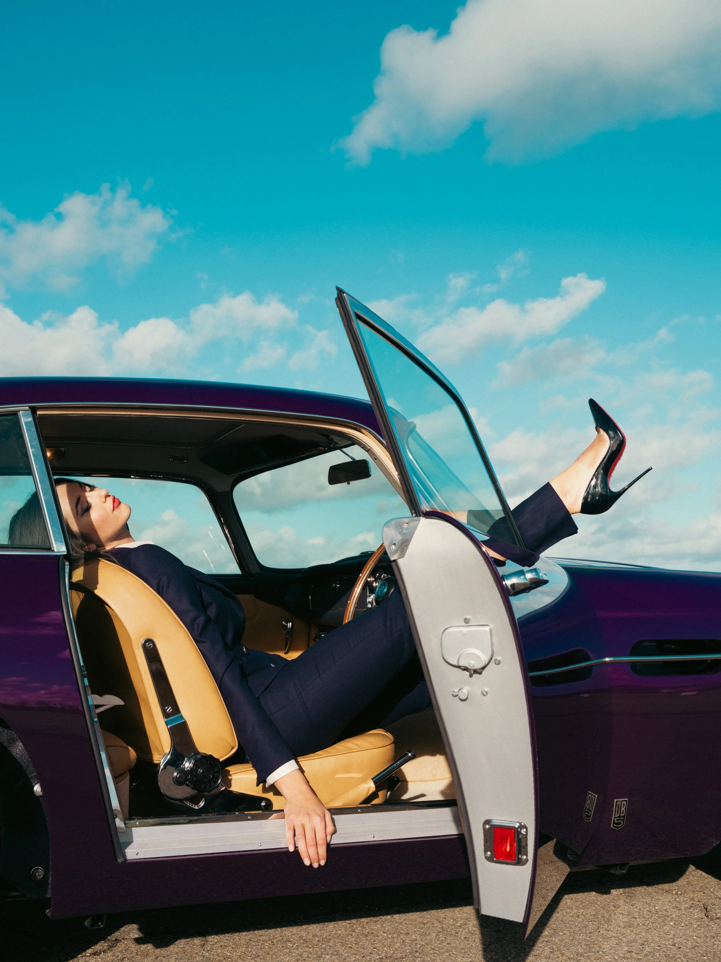 Woman wearing a navy women’s tailored wool suit with stiletto heels, reclining in a classic car for Paget London editorial