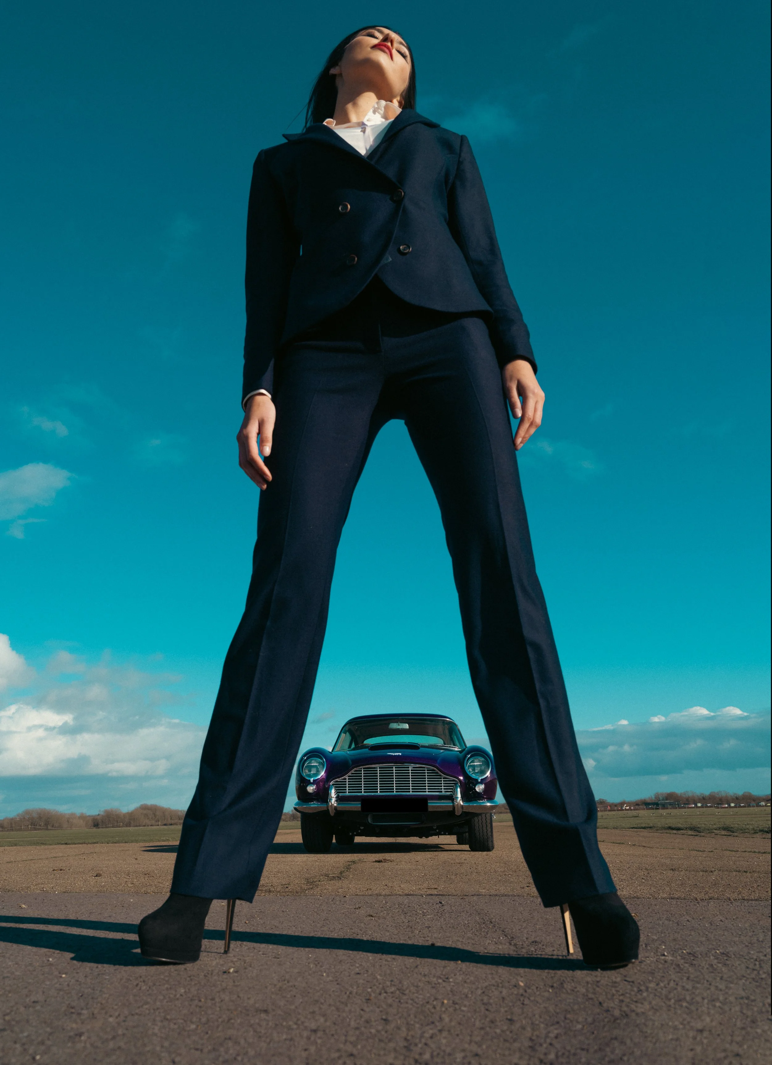 Woman wearing a navy women’s tailored wool trouser suit with straight-leg trousers and stiletto heels, photographed with a classic car for  Paget London editorial