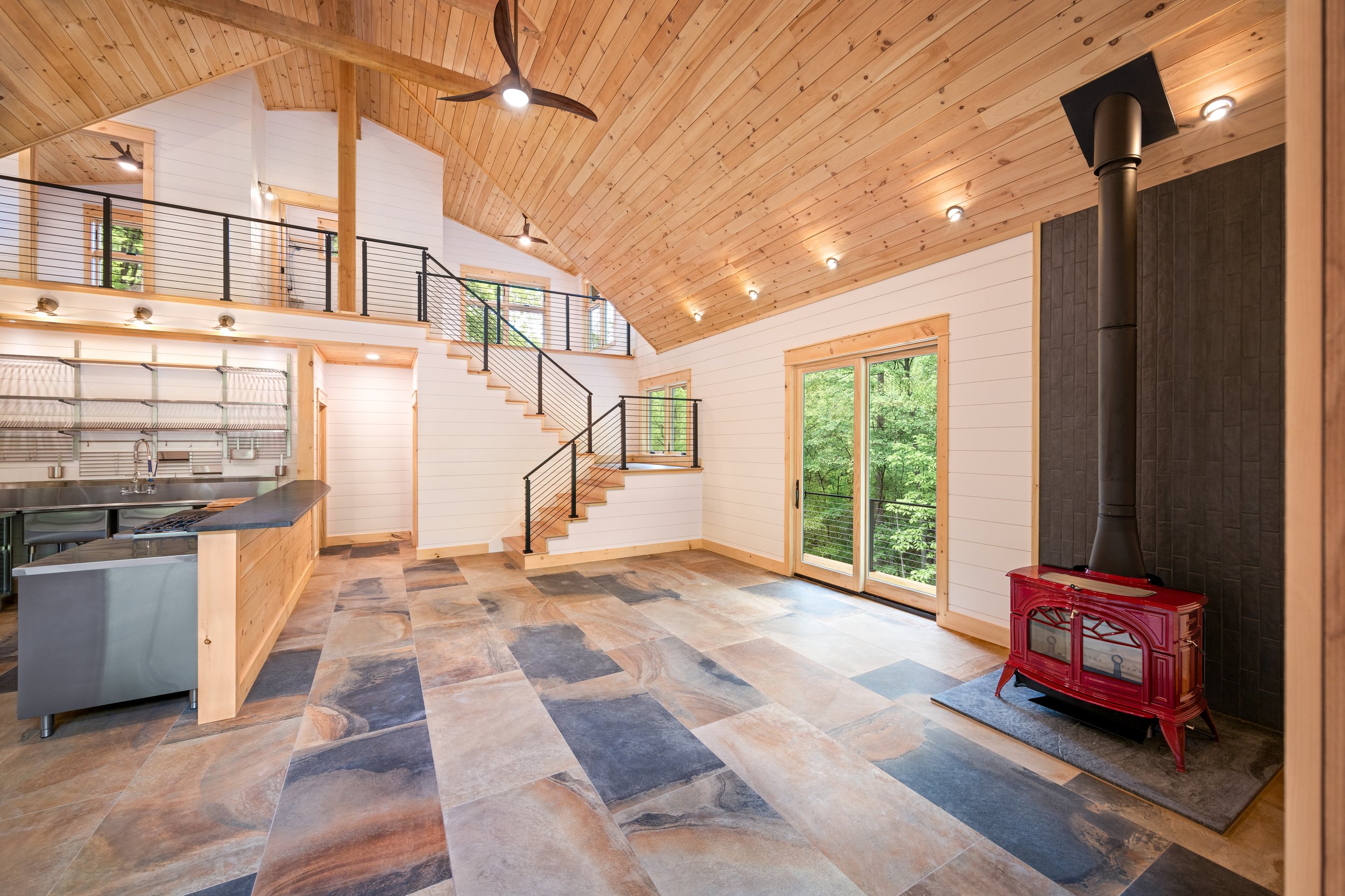 Open-concept great room featuring vaulted tongue-and-groove wood ceilings, a custom staircase with cable railing, large-format tile flooring, a modern wood stove, and expansive windows overlooking the surrounding forest in a custom blue ridge mountai