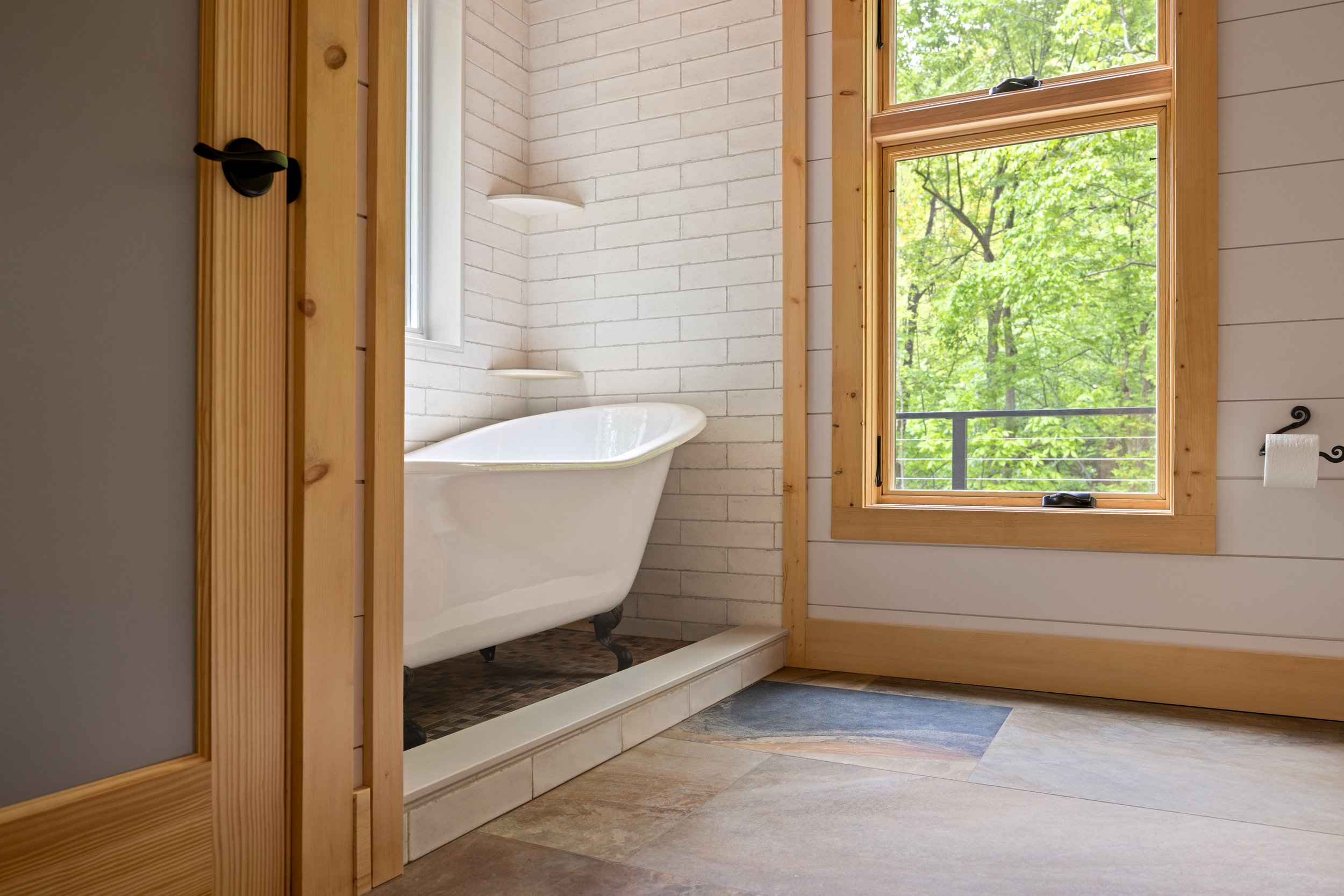 Primary Bathroom featuring a freestanding soaking tub, white subway tile surround, tongue-and-groove wood ceiling, large picture windows overlooking the forest, and natural wood trim in a custom mountain home in Nelson County, Virginia.