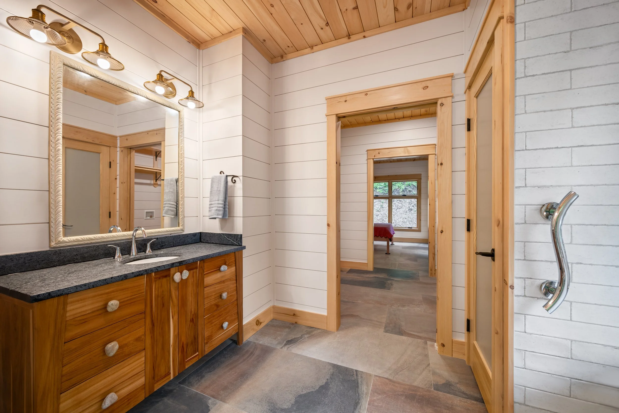 Nestled deep in the woods of Nelson County, VA. Custom bathroom featuring a natural wood vanity, granite countertop, white shiplap walls, tongue-and-groove wood ceiling, and large-format tile flooring.