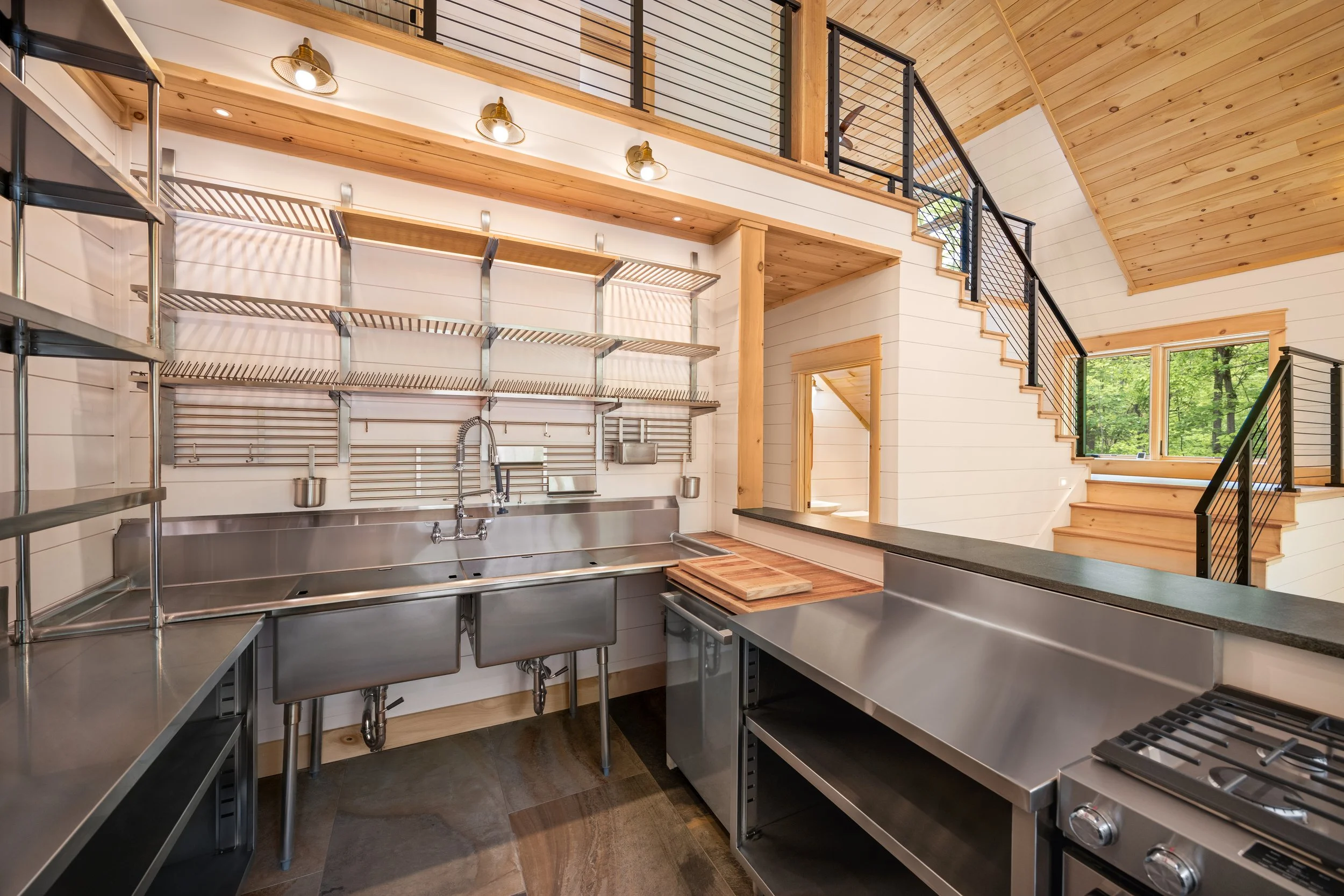 Commercial-style kitchen featuring stainless steel workstations, open shelving, a professional double-basin sink, custom prep island, and vaulted tongue-and-groove wood ceilings in a custom mountain home in Nelson County, Virginia.