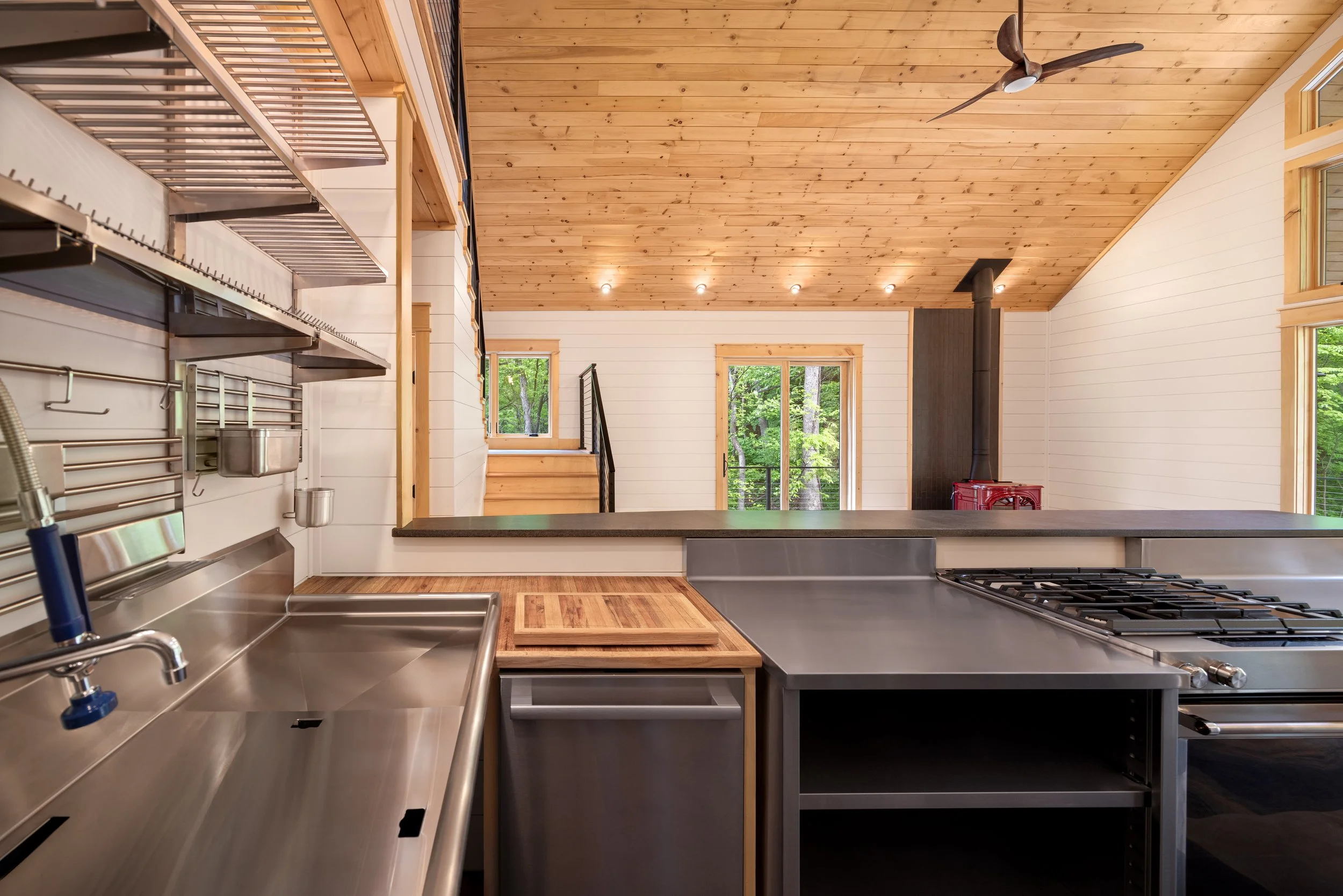 Commercial-style kitchen featuring stainless steel workstations, open shelving, a professional double-basin sink, custom prep island, and vaulted tongue-and-groove wood ceilings in a custom mountain home in Nelson County, Virginia.