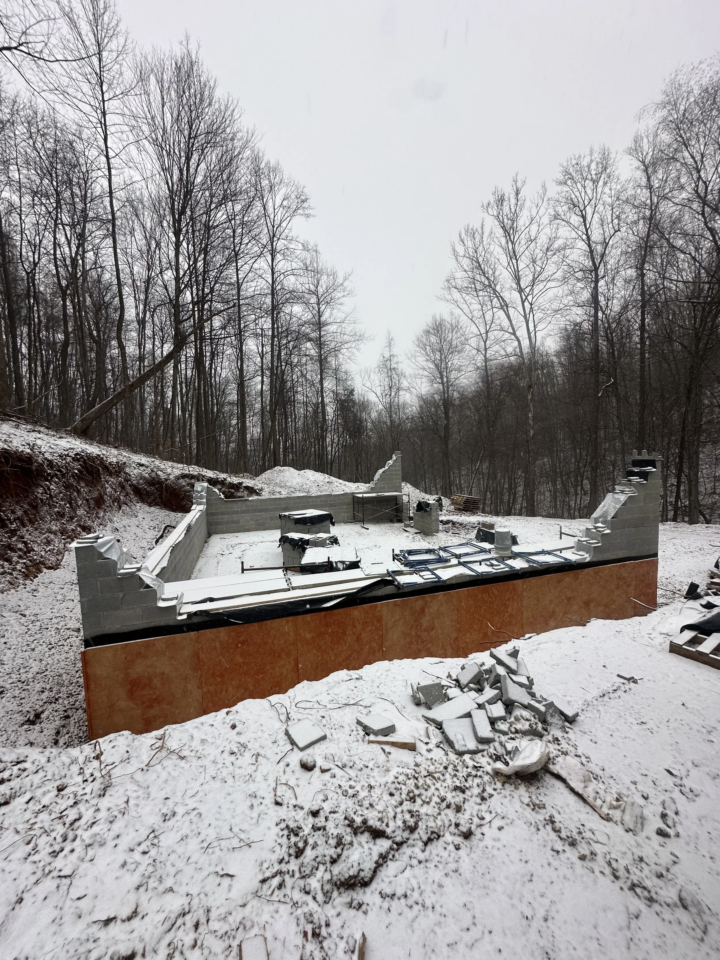 Construction site in a snowy forest showing the foundation of a building with cinder block walls and construction materials.