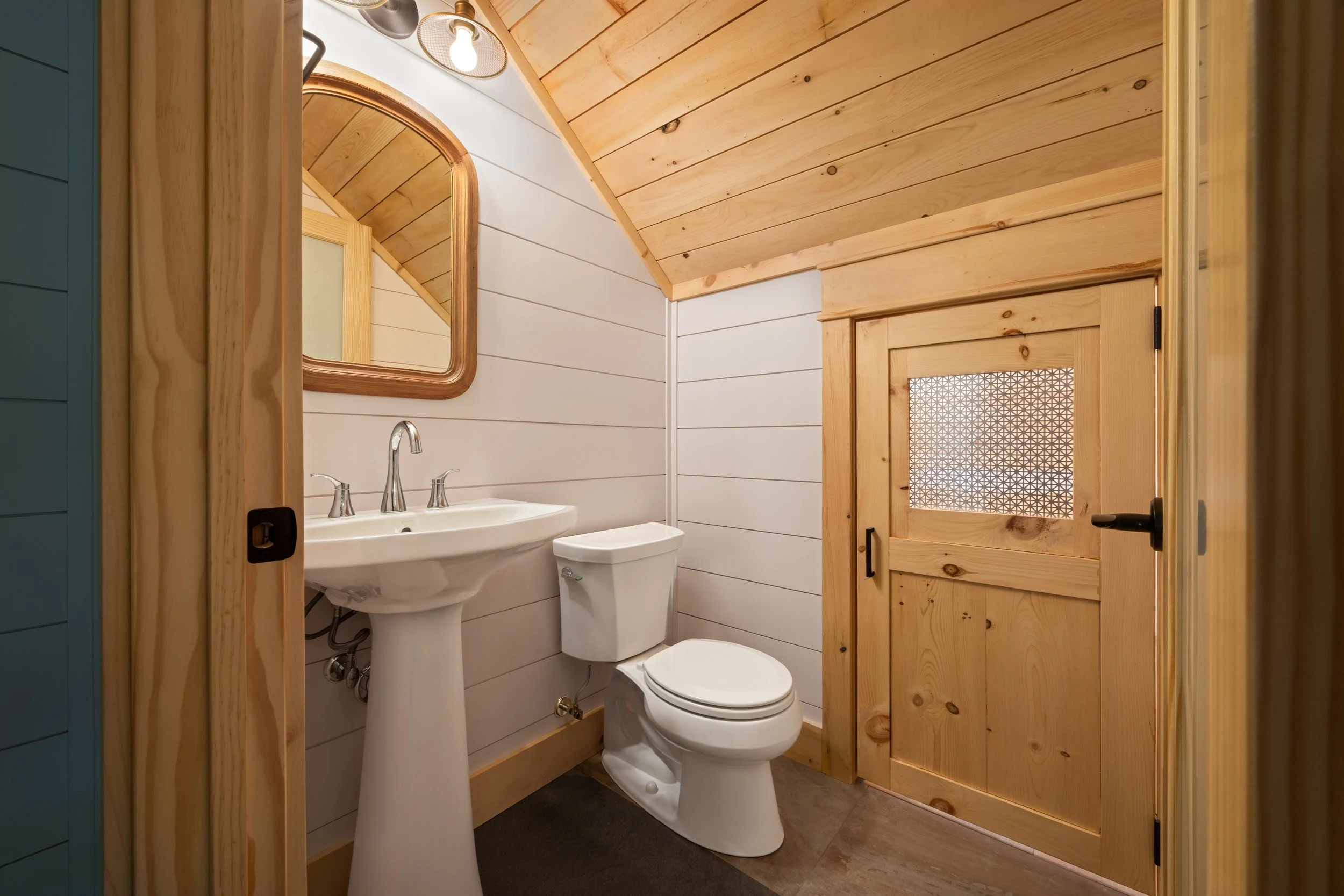 Half bathroom featuring white shiplap walls, a tongue-and-groove wood ceiling, natural wood trim, a pedestal sink, and a custom wood barn-style door with decorative privacy glass in a custom mountain home in Nelson County, Virginia.