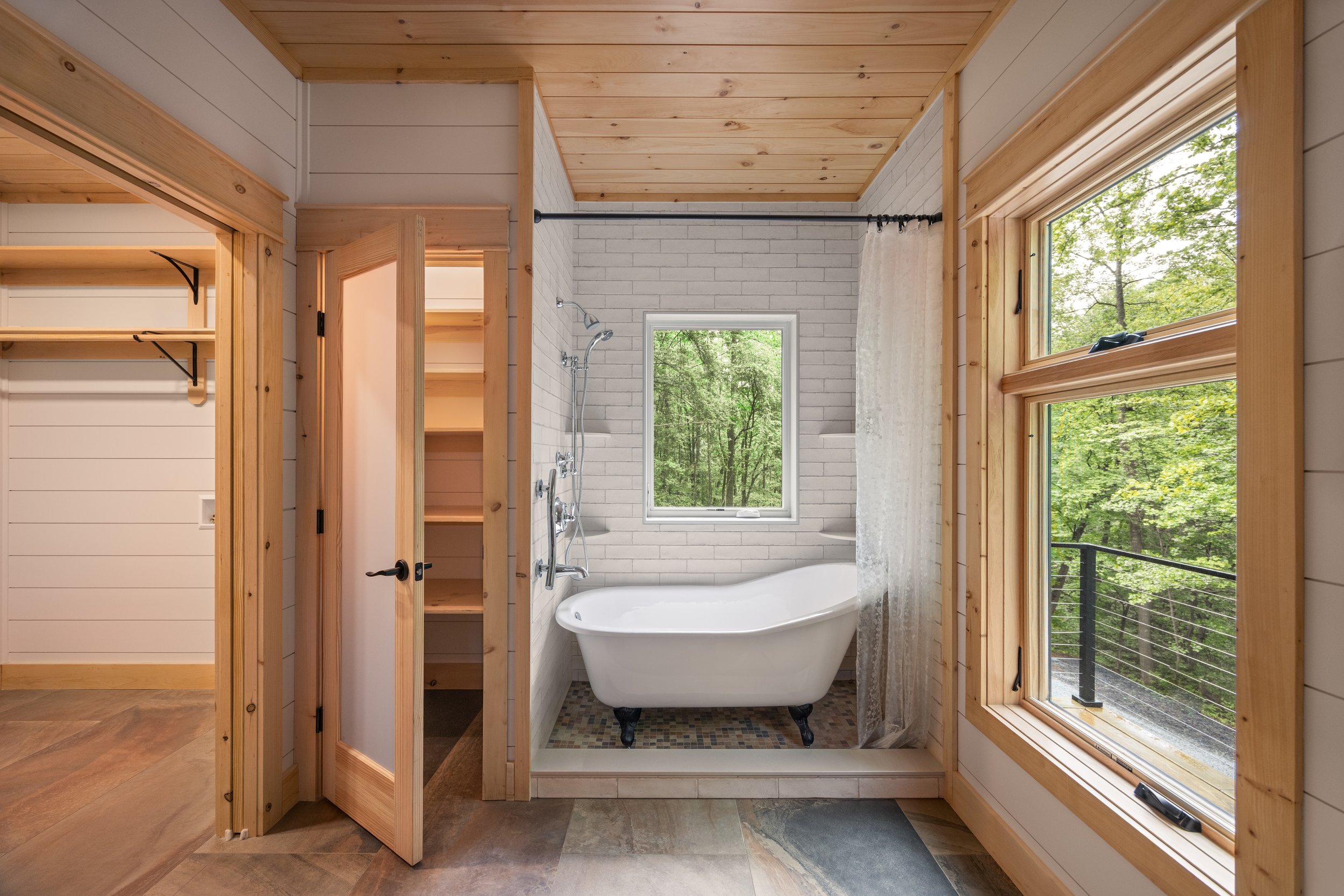 Primary Bathroom featuring a freestanding soaking tub, white subway tile surround, tongue-and-groove wood ceiling, large picture windows overlooking the forest, and natural wood trim in a custom mountain home in Nelson County, Virginia.