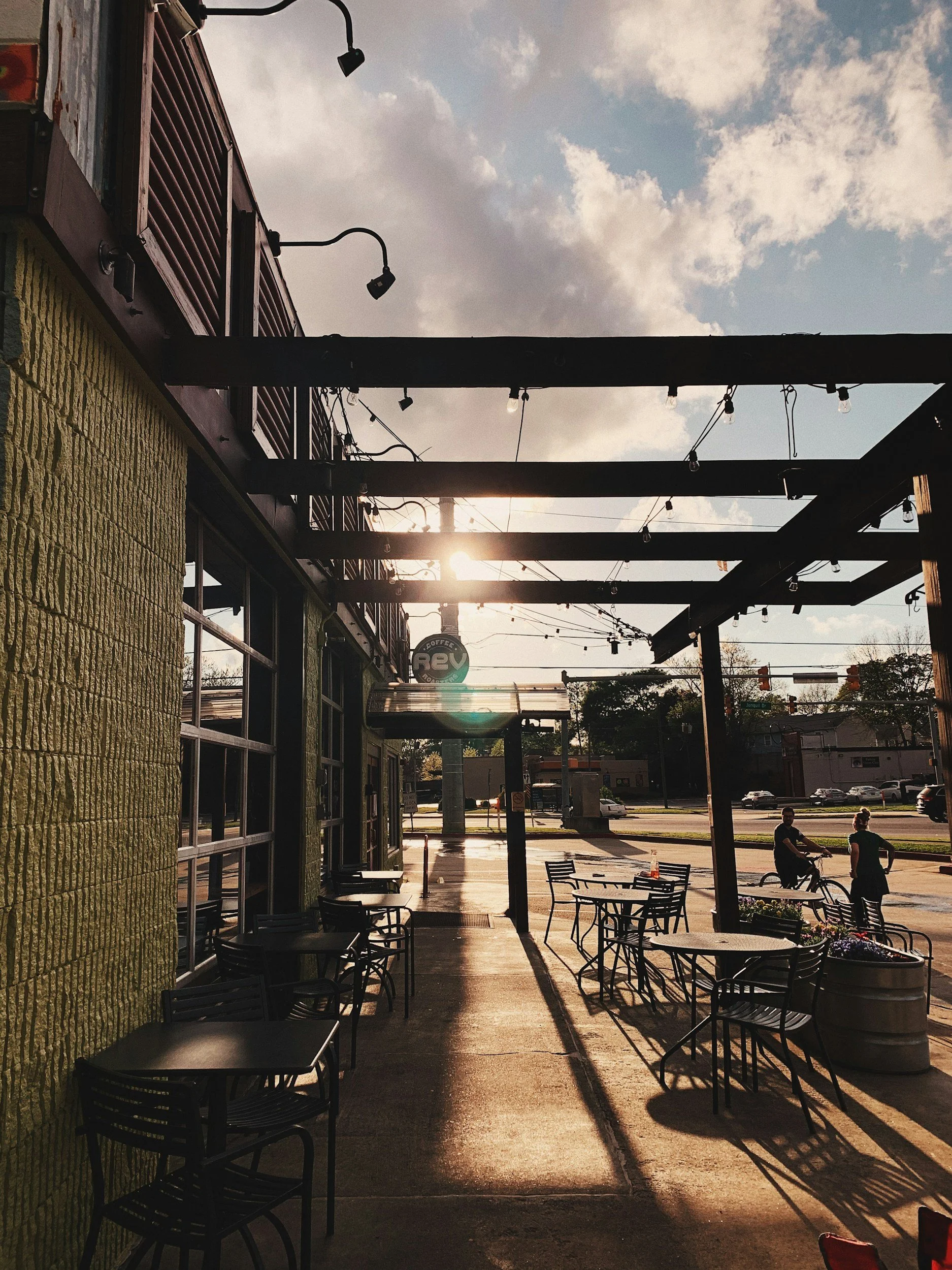 Empty outdoor patio of a cafe with black tables and chairs, string lights, and a green painted brick wall, with people and cars in the background during sunset.