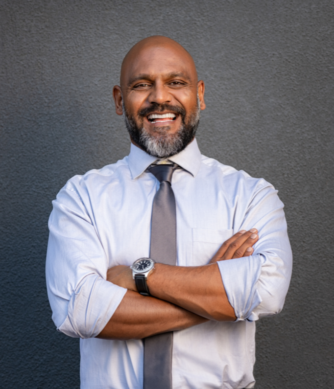 A smiling middle-aged man with a beard and bald head, wearing a light blue dress shirt, a grey tie, and a wristwatch, standing with arms crossed against a dark grey wall background.