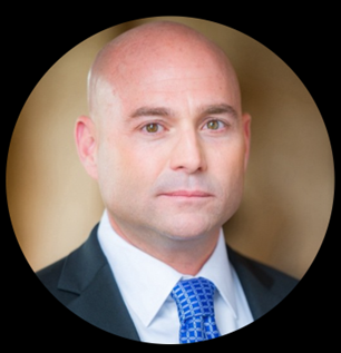 A bald man in a suit and blue tie posing for a portrait against a neutral background.