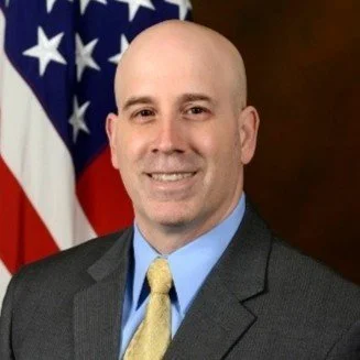 A bald man in a gray suit with a blue shirt and yellow tie, smiling, with the American flag in the background.