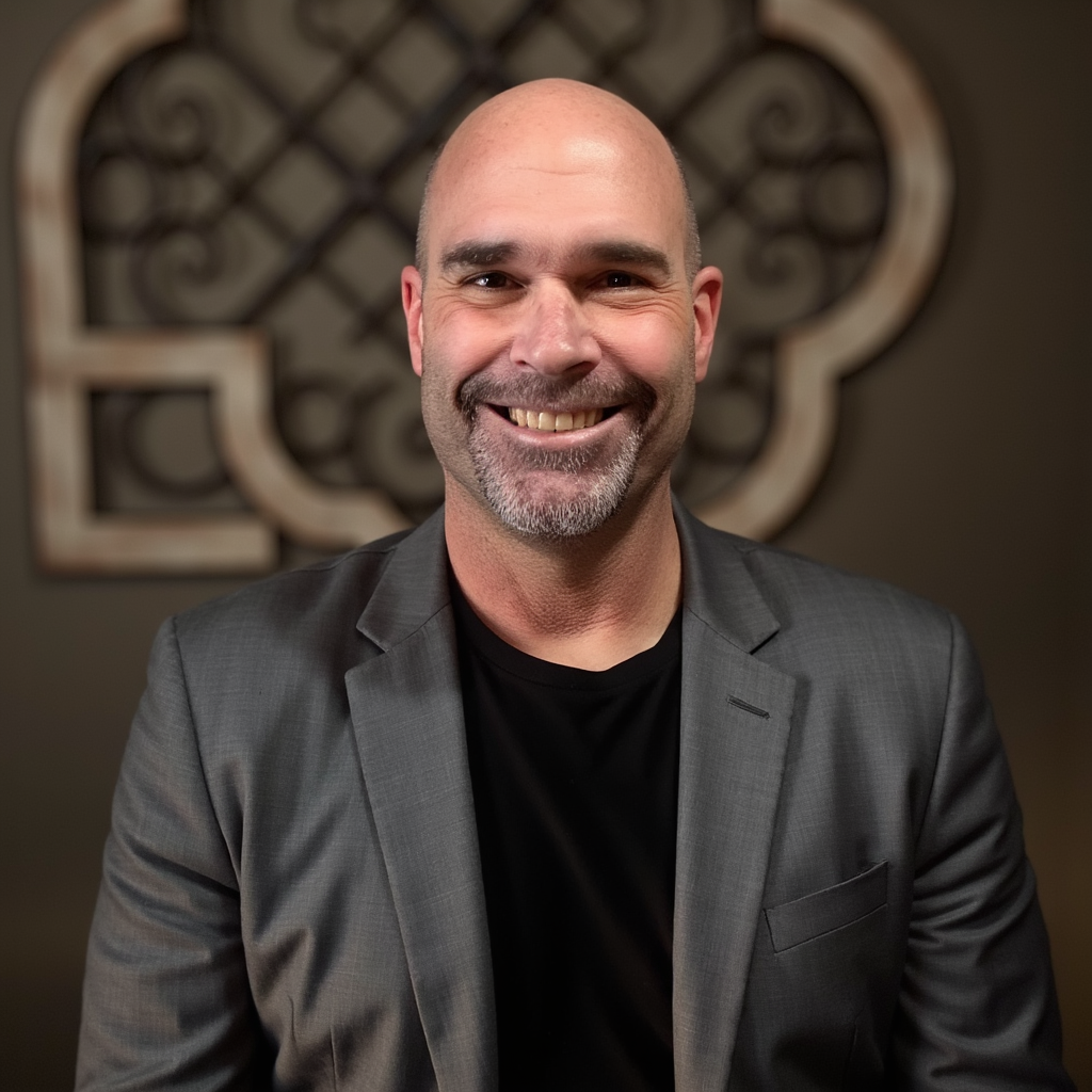 A smiling man with a shaved head and gray beard, wearing a gray suit and black shirt, standing in front of an ornate decorative wall piece.