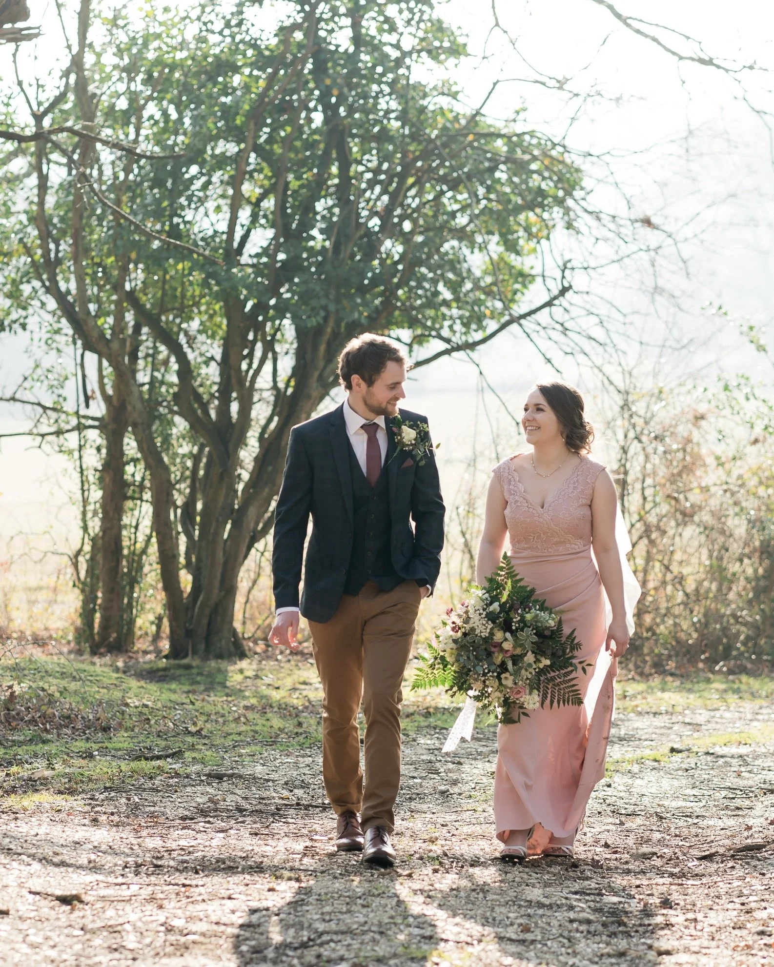 Newlywed couple walking outdoors. Groom is dressed in brown chinos with a dark blue jacket and waitcoat, and the bride has a pibk wedding dress and is carrying a large bouquet.